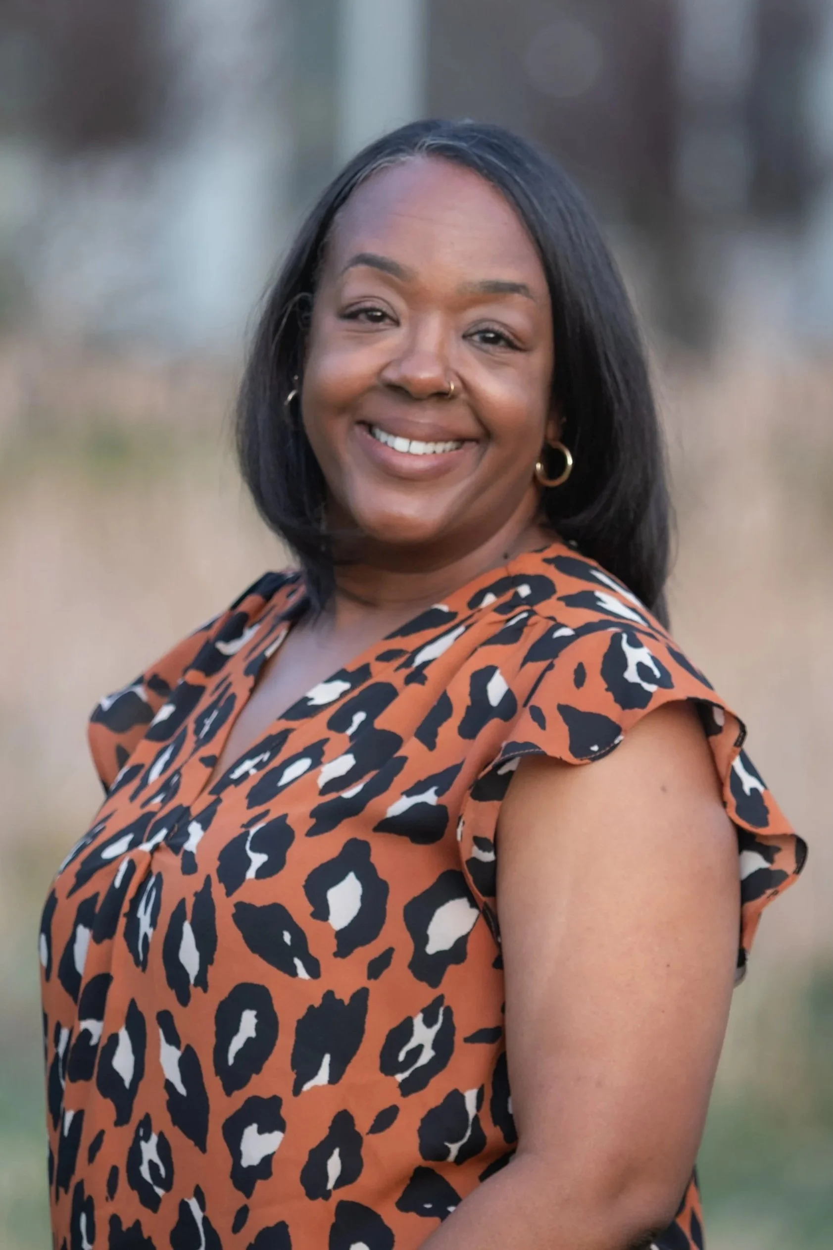 A woman smiling outdoors, wearing a sleeveless animal print dress with ruffled shoulders and gold hoop earrings.