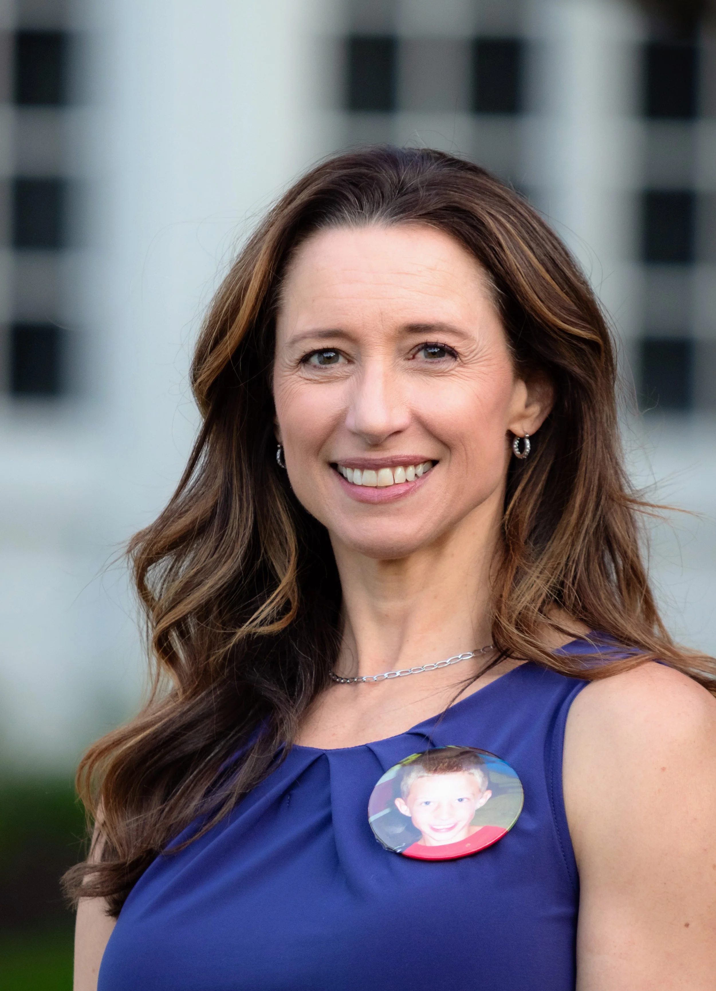 A woman with long brown hair wearing a blue dress, silver jewelry, and a button with a child's photo, standing outdoors with a building in the background.