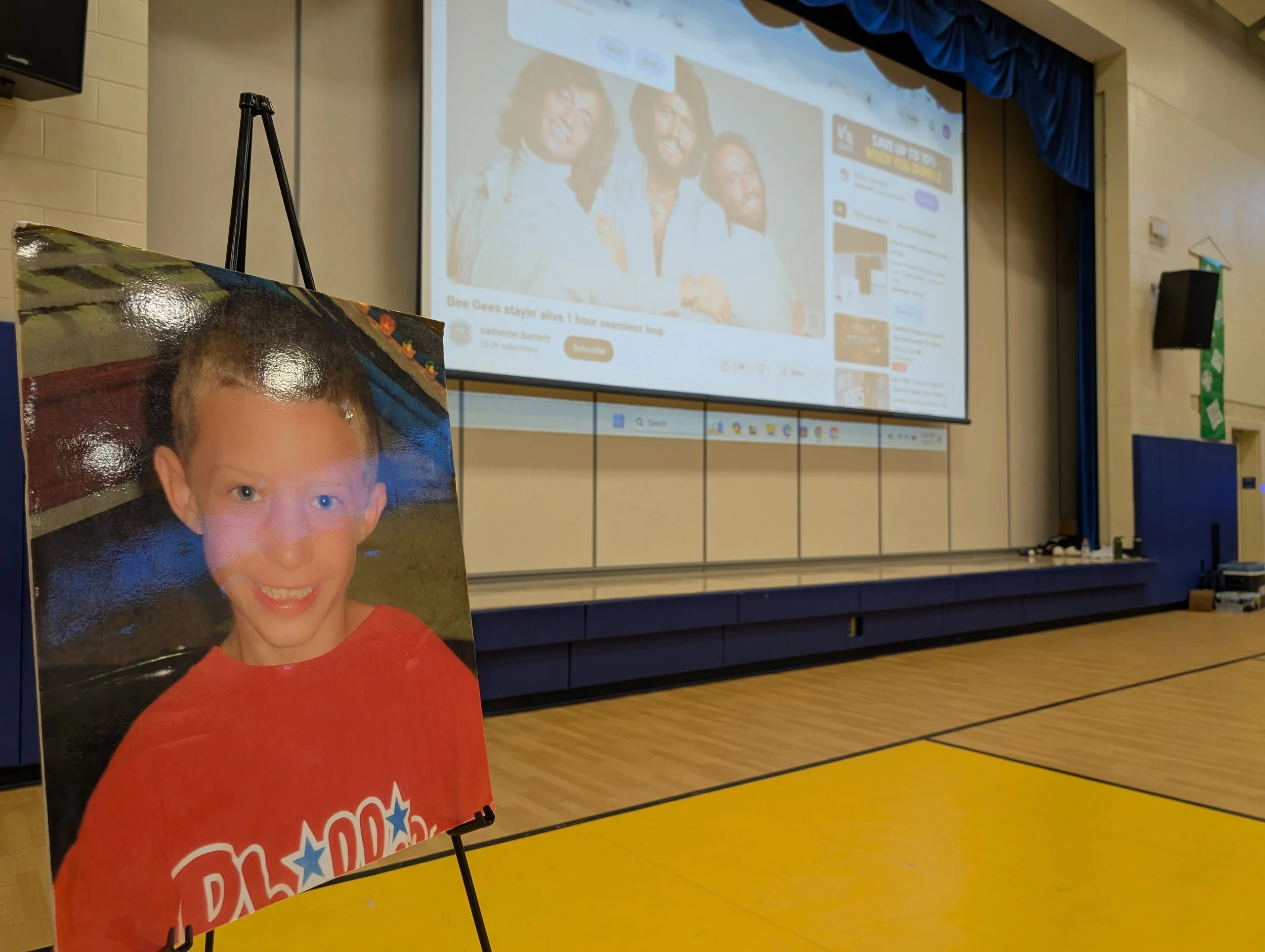 A framed photo of a young boy with short light brown hair, smiling, wearing a red shirt with text and stars. The photo is placed on an easel in front of a school gymnasium or auditorium stage with a large screen displaying a YouTube video.
