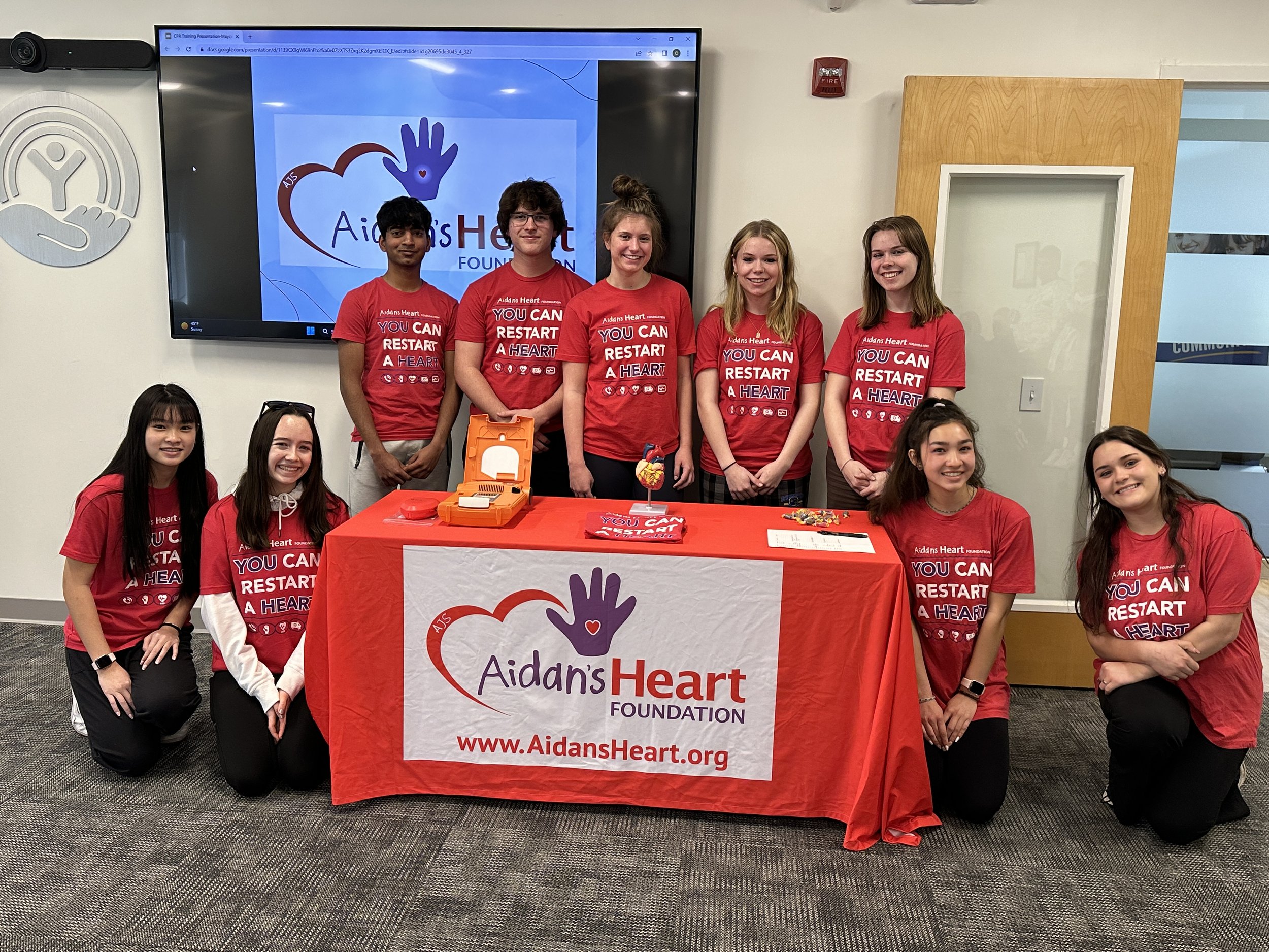 Group of nine young people, wearing matching red shirts with 'You Can Restart a Heart' slogan, gathered around a table with an AED device and materials for a heart health awareness event, in a room with a TV screen displaying the AidansHeart Foundation logo.