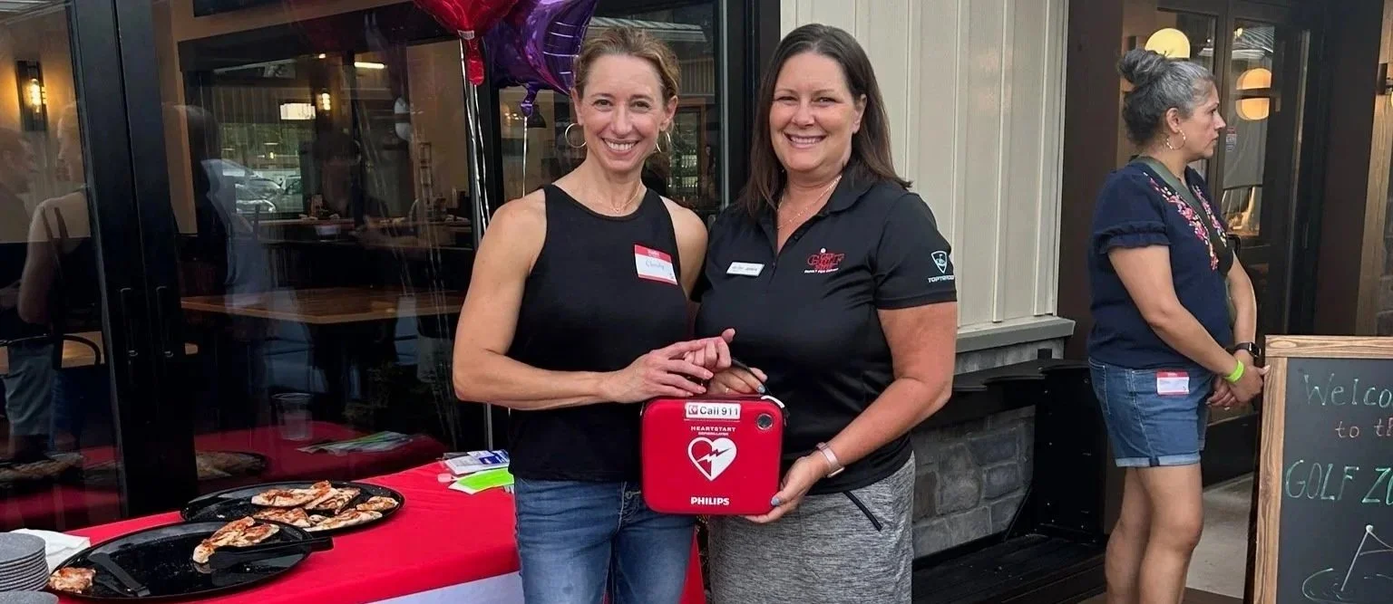 Two women standing outside a restaurant, smiling and holding a red automated external defibrillator (AED). One woman has short blonde hair and is wearing a black sleeveless top, while the other has shoulder-length brunette hair and is dressed in a black polo shirt and gray pants. Behind them is a table with pizza and purple balloons, and to the right, a woman with gray hair in a bun wearing a navy blue shirt and denim shorts stands near a chalkboard sign.