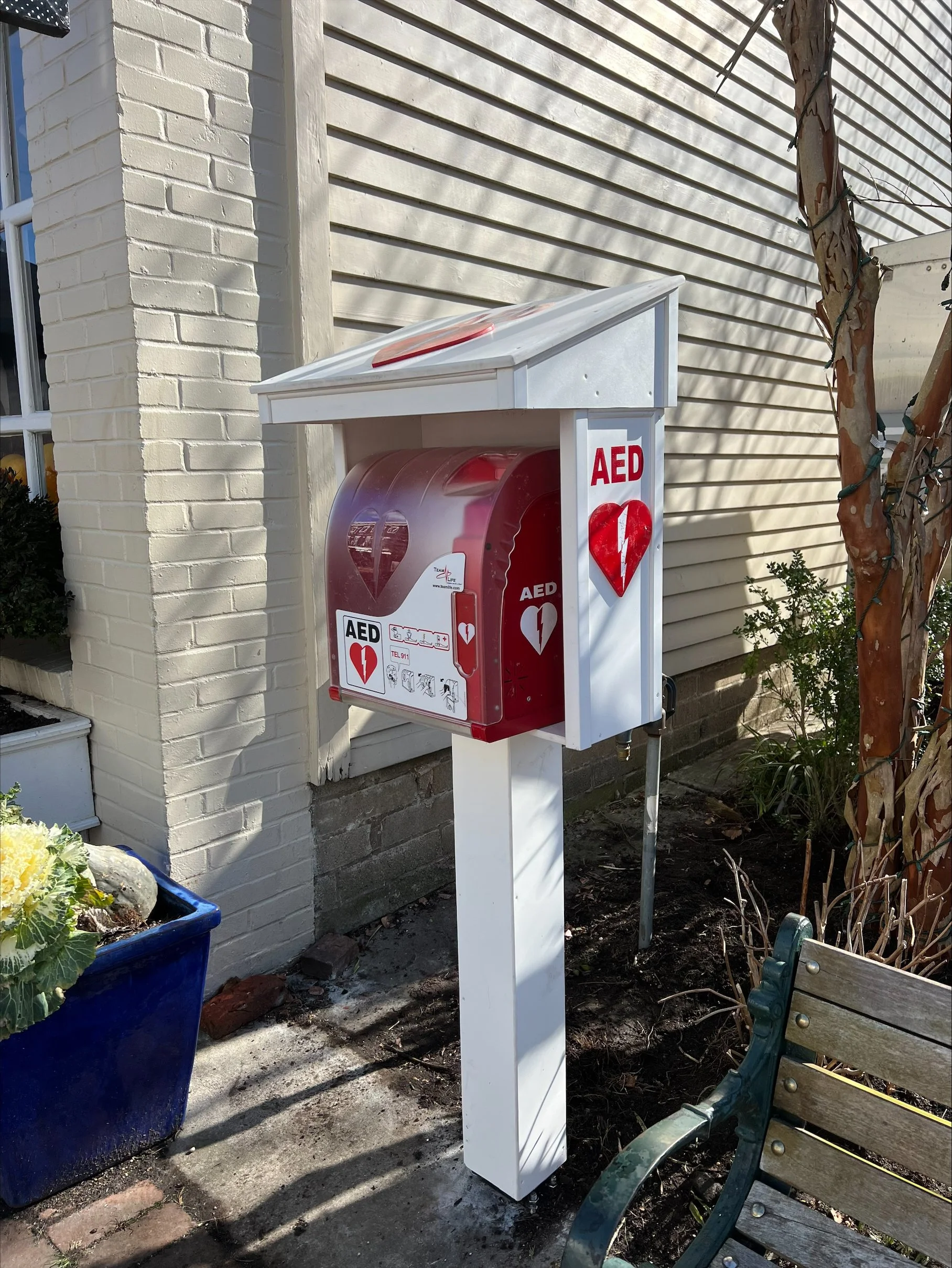 Red automated external defibrillator (AED) station outside next to a white brick wall, with a white house siding behind. There is a blue flowerpot with yellow and green plants on the left and a wooden park bench on the right. The AED is within a protective white and red box with a heart symbol and medical instructions.