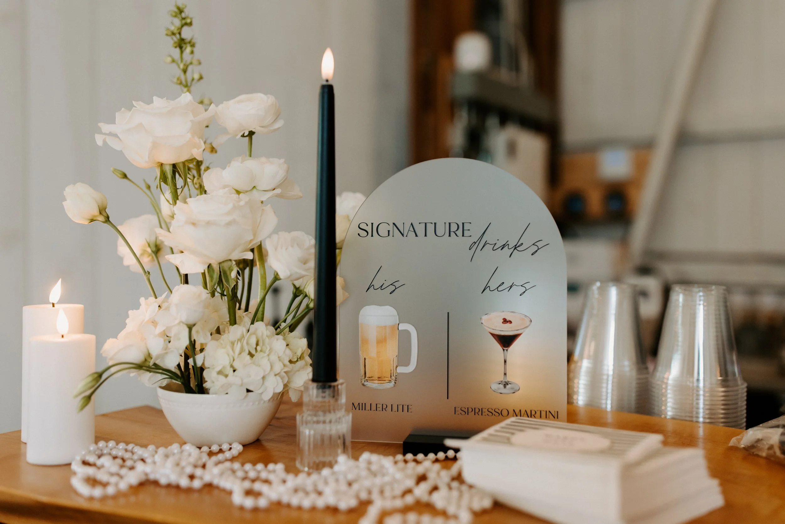 Table setup with white flowers, lit candles, a sign for signature drinks showing Miller Lite and Espresso Martini, cups, and a pearl necklace.