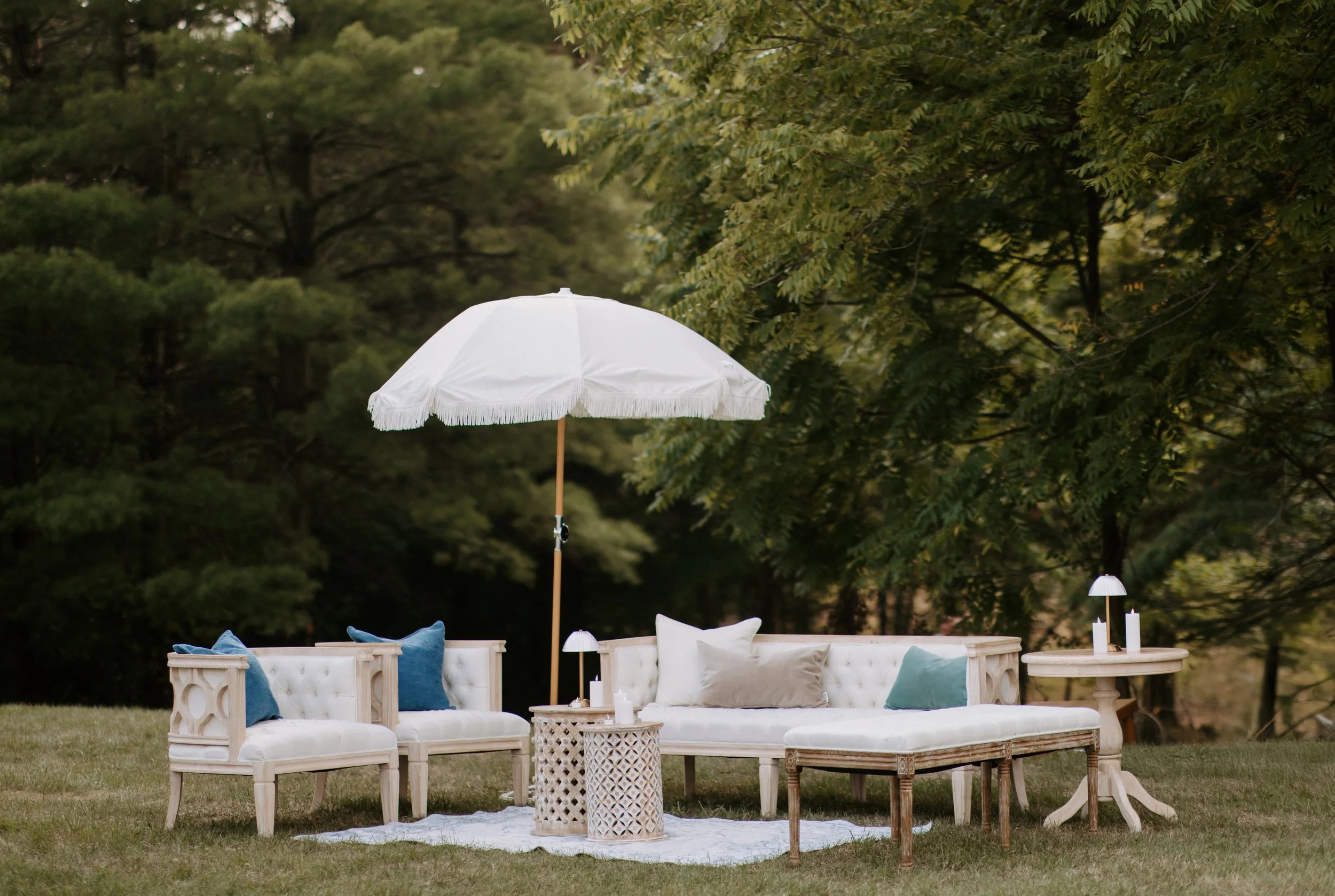 Outdoor seating area with a white umbrella, two sofas with white cushions and throw pillows, and small tables with lamps, set on a grassy yard amidst trees.