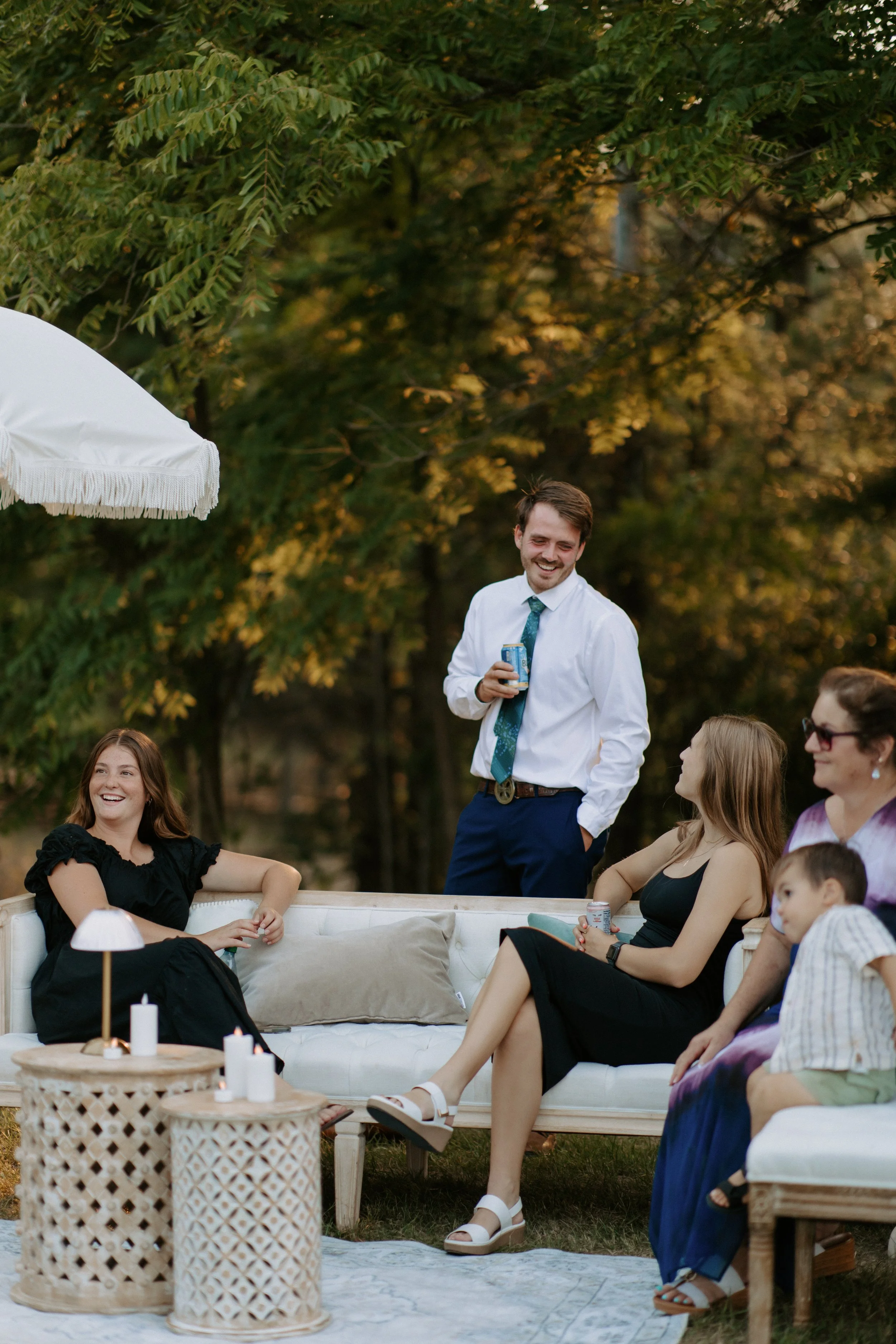 Group of people socializing outdoors in a garden or park, sitting on white furniture with candles on side tables, trees in the background, a man in a white shirt holding a can, and others smiling and chatting.