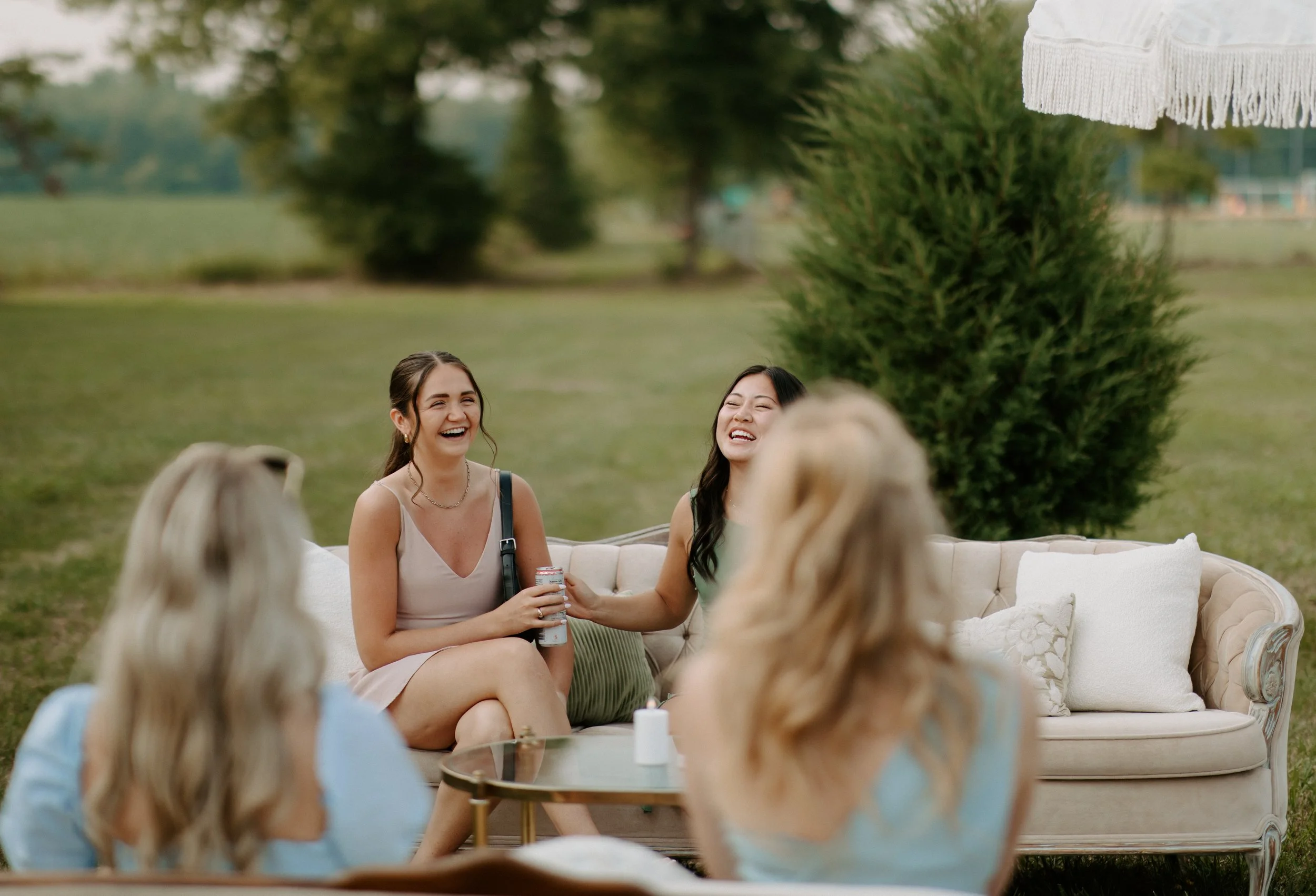 A group of four women enjoying a social gathering outdoors in a park, sitting on a vintage couch and chairs, laughing and smiling.