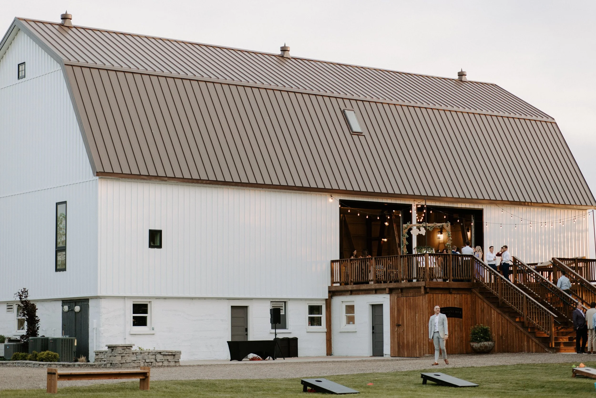 An updated rustic barn and wooden deck hosting an event, featuring string lights and guests on the staircase, surrounded by a lawn with cornhole game boards.