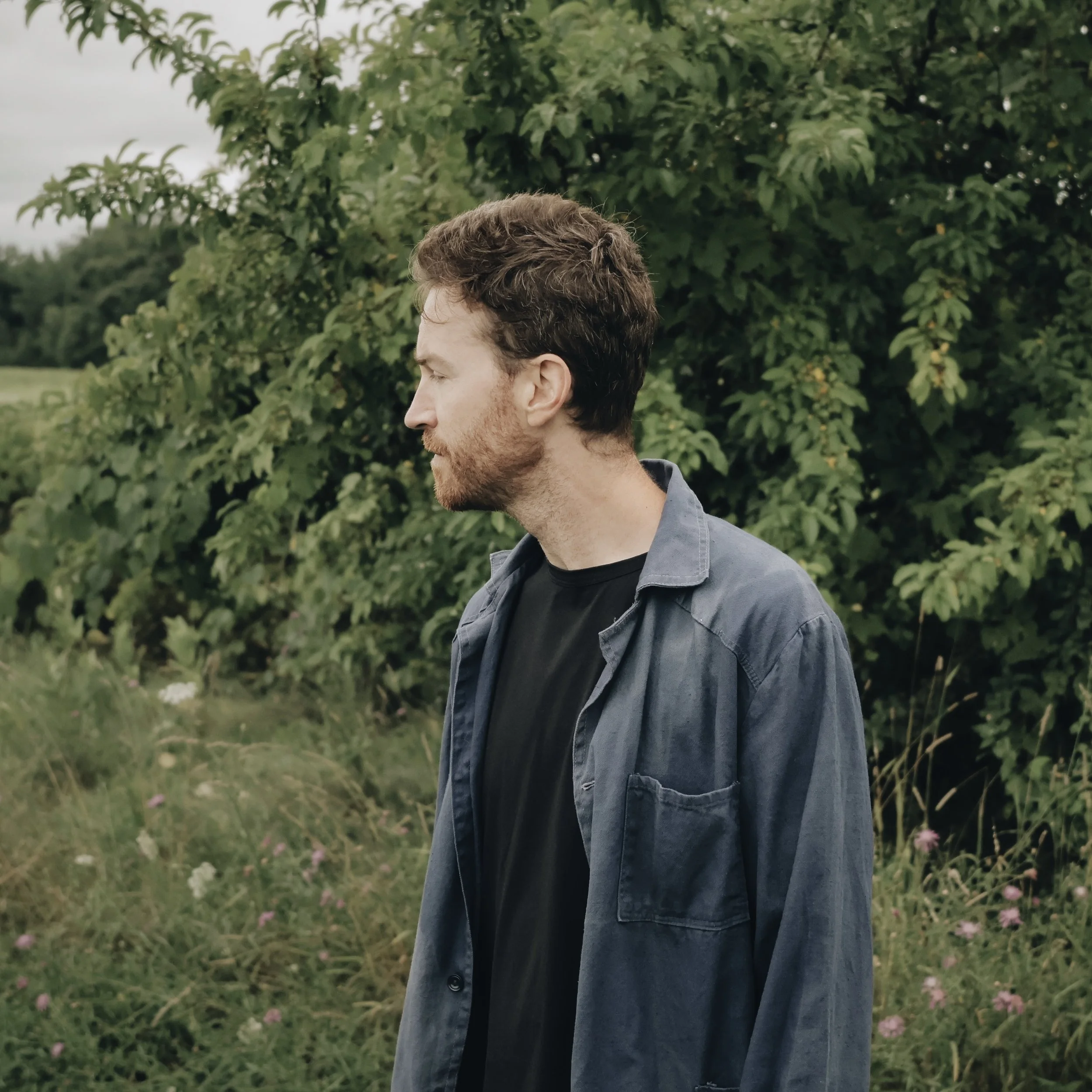 A man with brown hair and a beard stands outdoors in front of dense green foliage, facing left with eyes closed.
