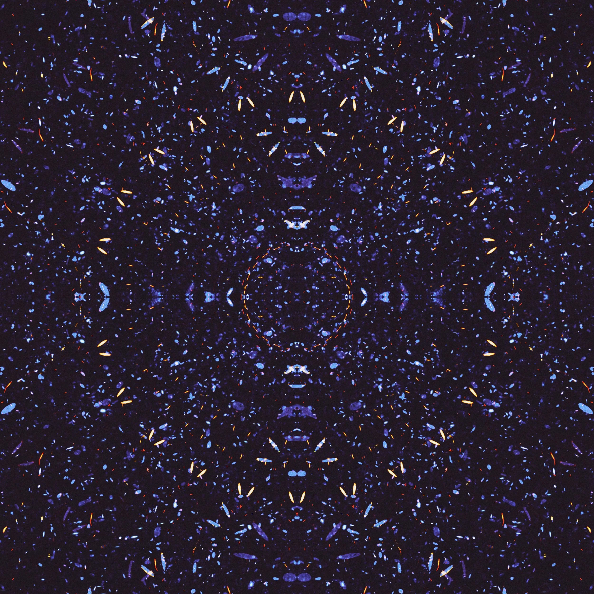 A symmetrical, abstract pattern of tiny, glowing, colorful particles on a dark background, resembling a kaleidoscope or a digital design.