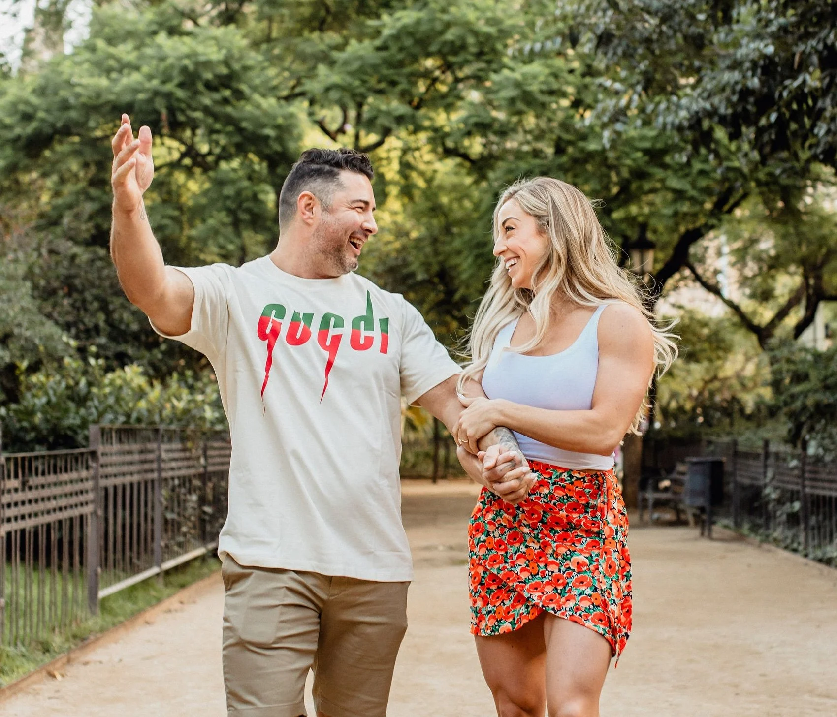 A young couple laughing and holding hands, walking in a park with trees and a pathway.