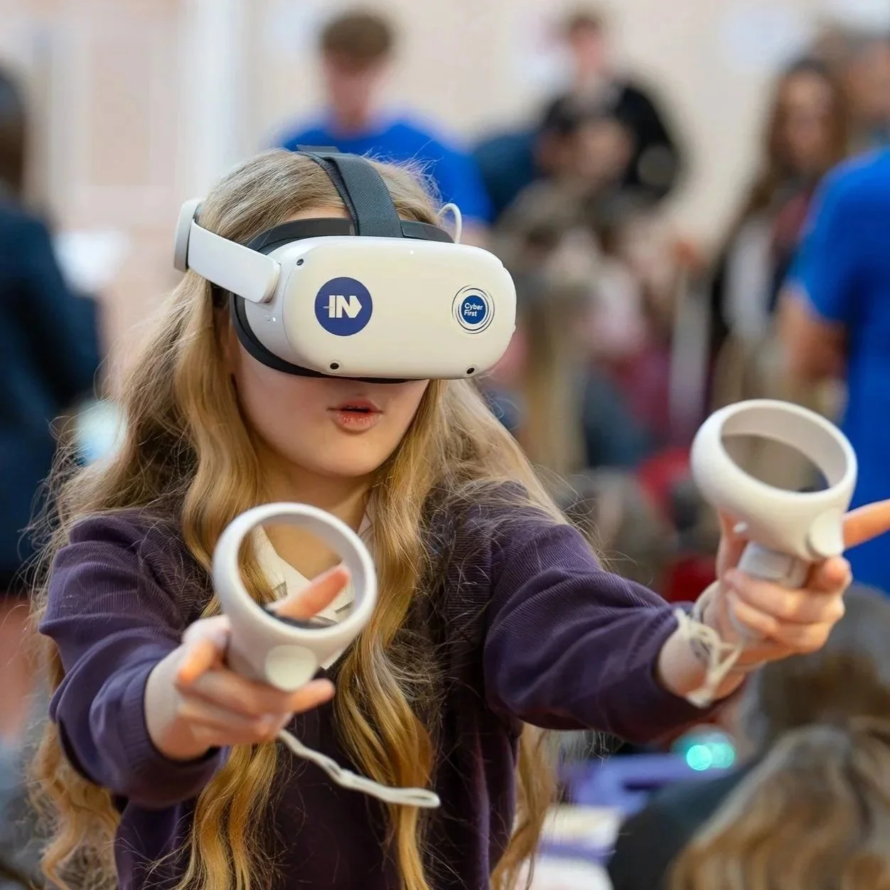 A young girl with long, curly blonde hair wearing a virtual reality headset and holding motion controllers, participating in a VR experience at a crowded indoor event.