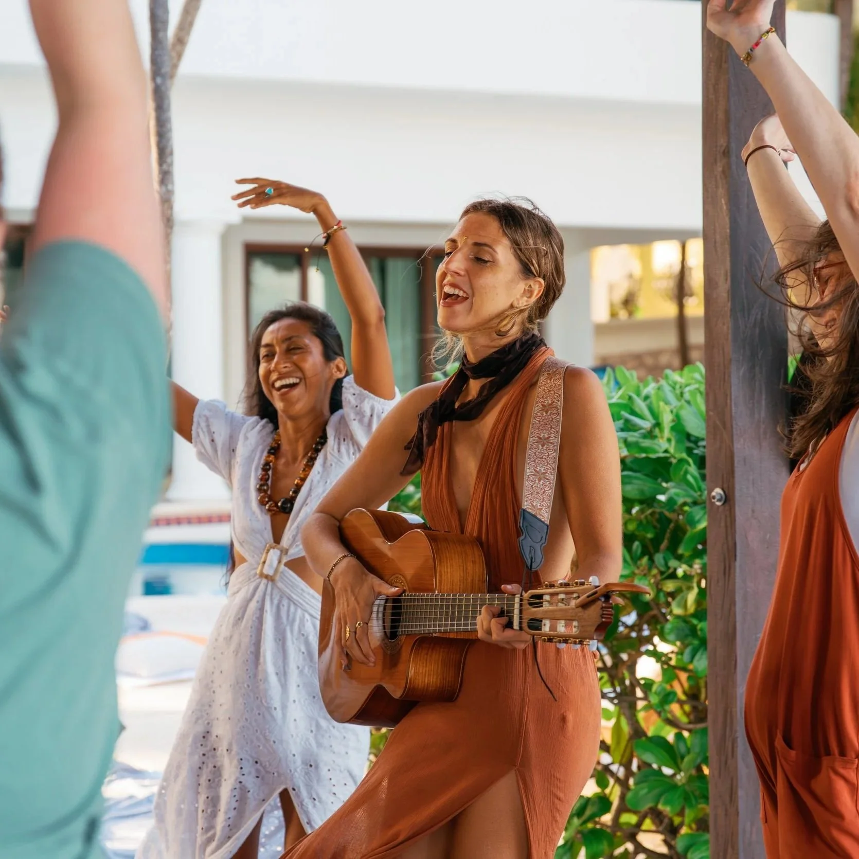 Three women are singing and playing guitar outdoors, possibly at a festive or social gathering, with greenery and a building in the background.