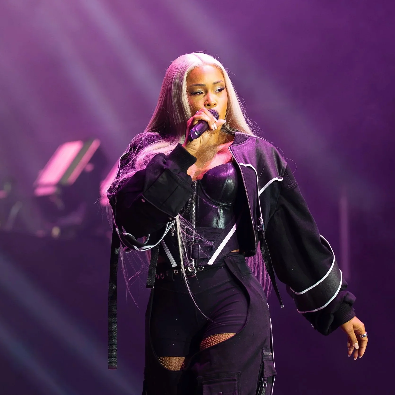 A female singer on stage, holding a microphone with purple lighting in the background, wearing a black jacket with white stripes and fishnet stockings.