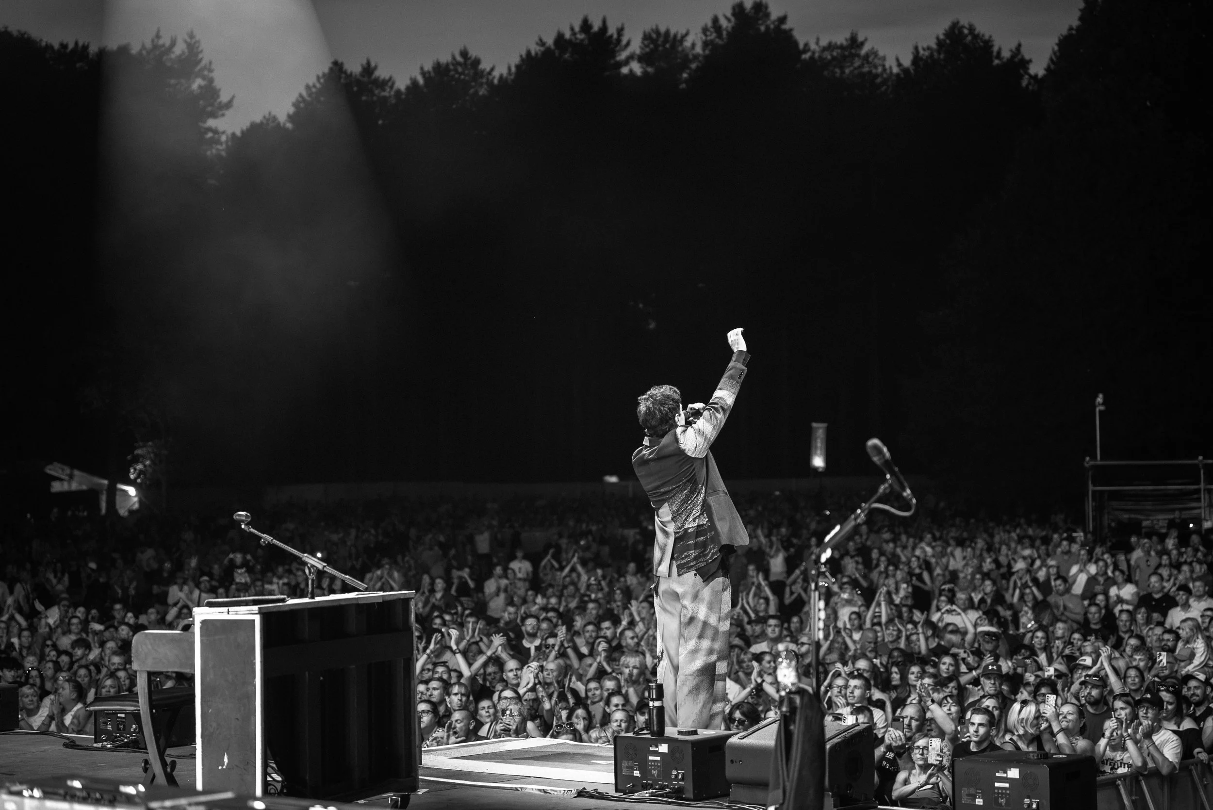 A performer on stage with a raised fist addressing a large seated audience outdoors at night, with trees in the background.
