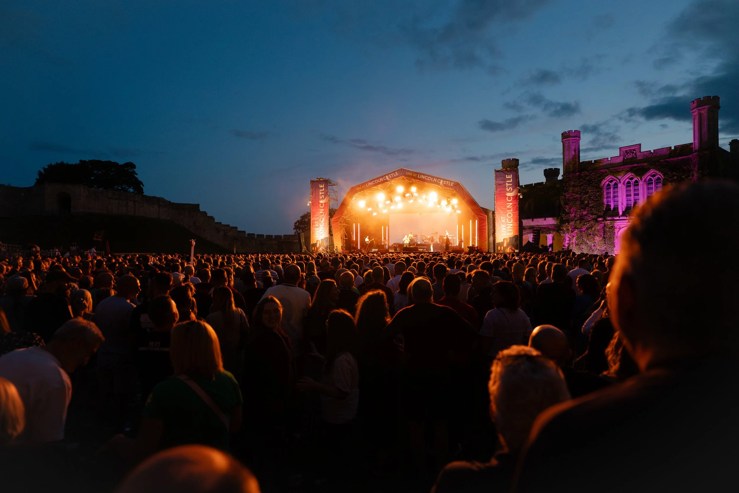 Outdoor concert at dusk with a large crowd watching a stage with bright lights. Castle-like structures are visible in the background, illuminated in purple and pink hues.