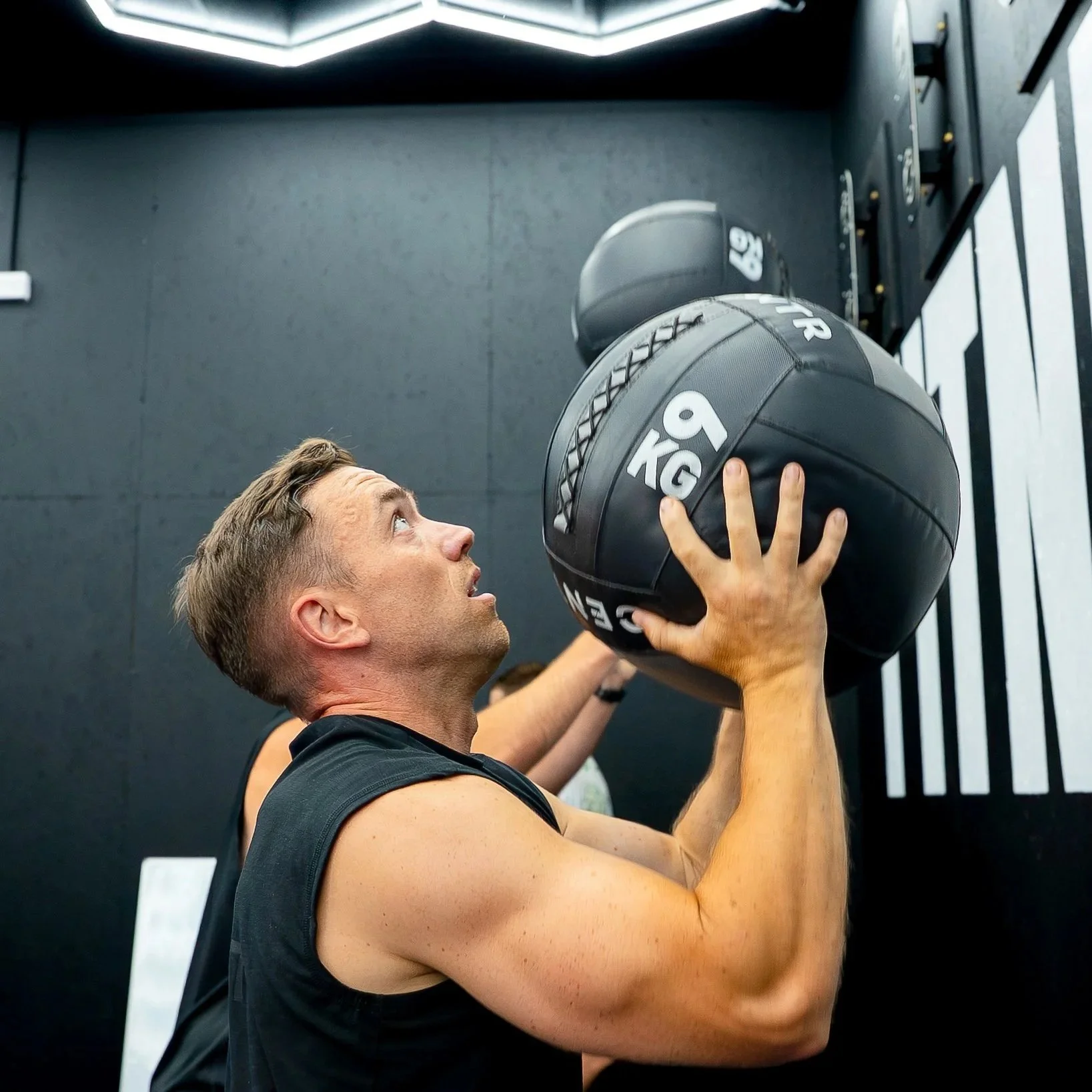 A man doing overhead shoulder presses with a black medicine ball in a gym.