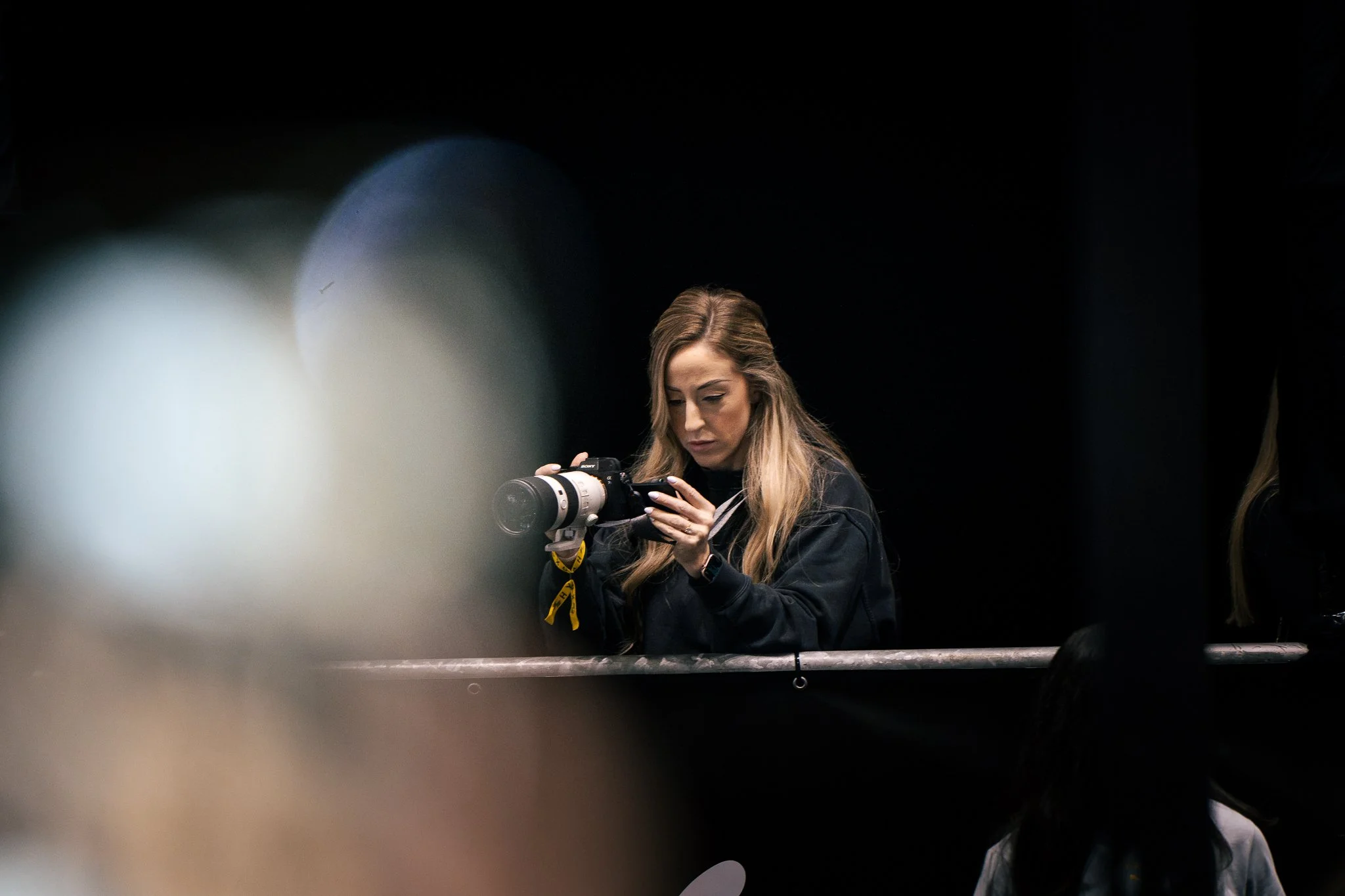 A woman with long hair, holding a professional camera, is standing behind a barrier in a dark setting, possibly during a photography or event setup.