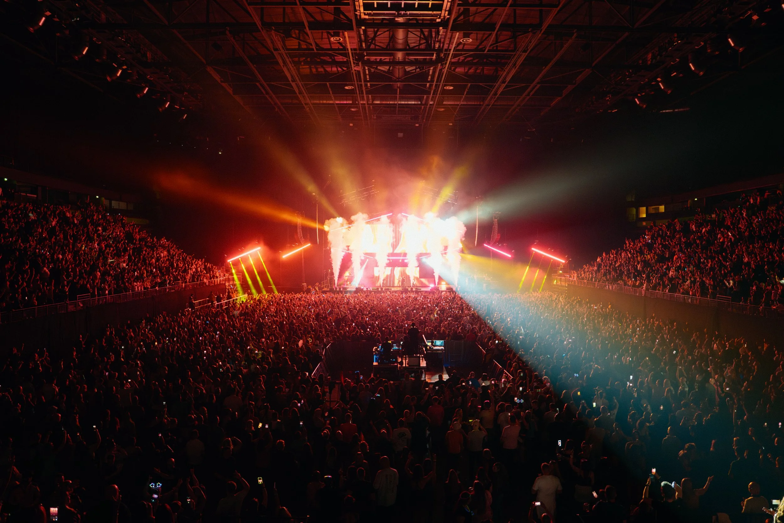 Large indoor concert with a crowd, bright stage lights, and smoke effects in a stadium or arena.