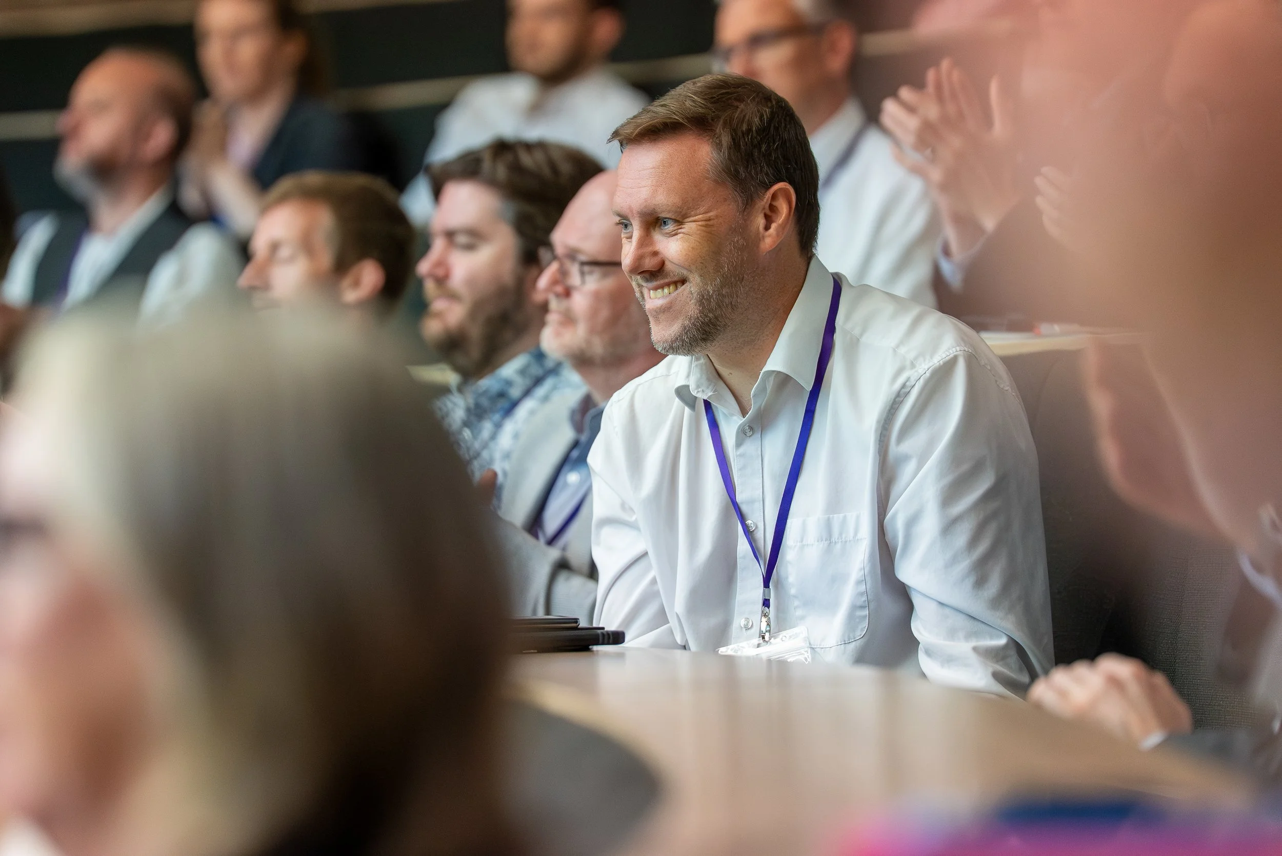 Smiling man with short brown hair and beard in white shirt at conference, surrounded by blurred audience members.
