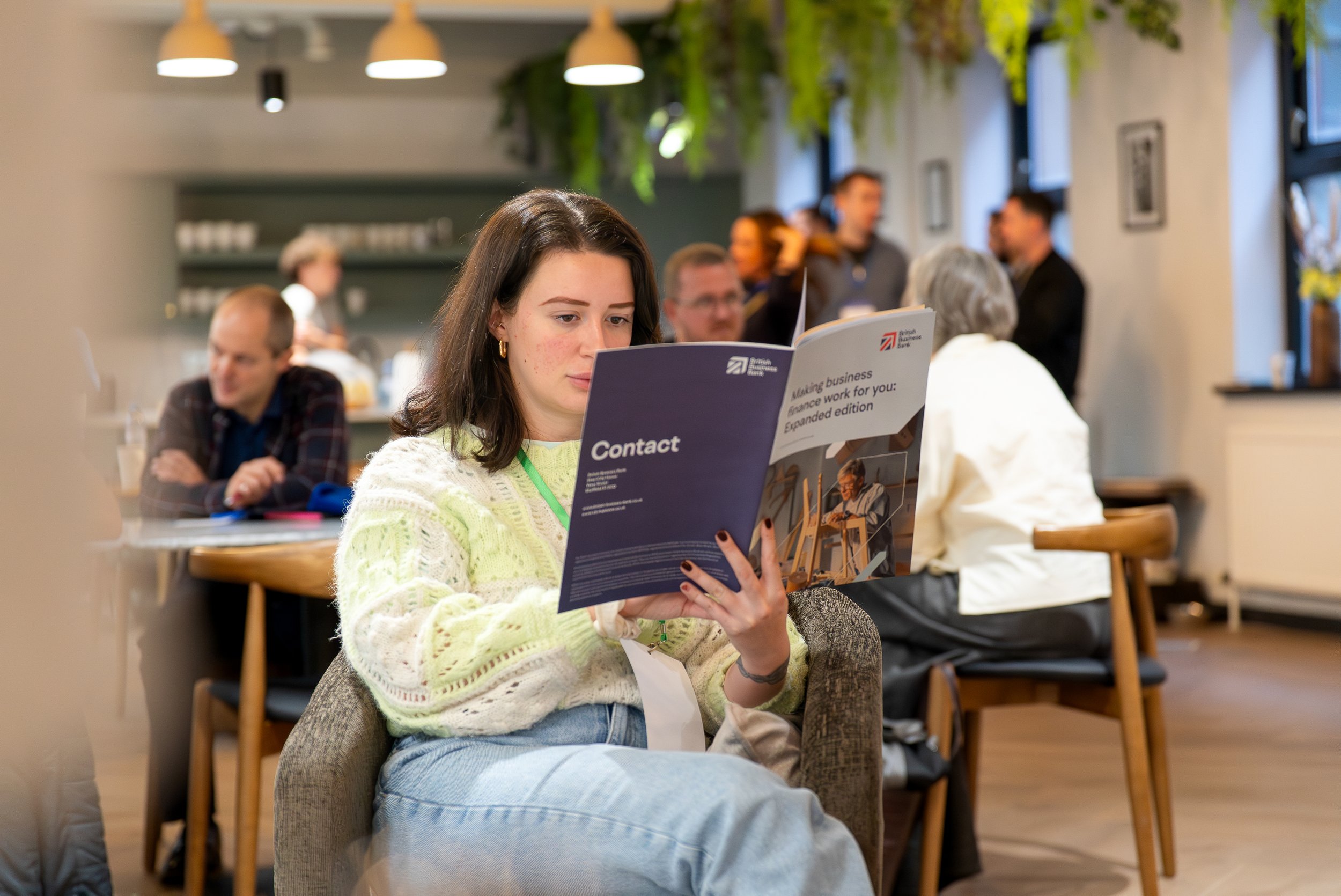 A young woman with dark hair and earrings reads a brochure at a busy indoor event, surrounded by other people sitting and standing, with green plants hanging from the ceiling.
