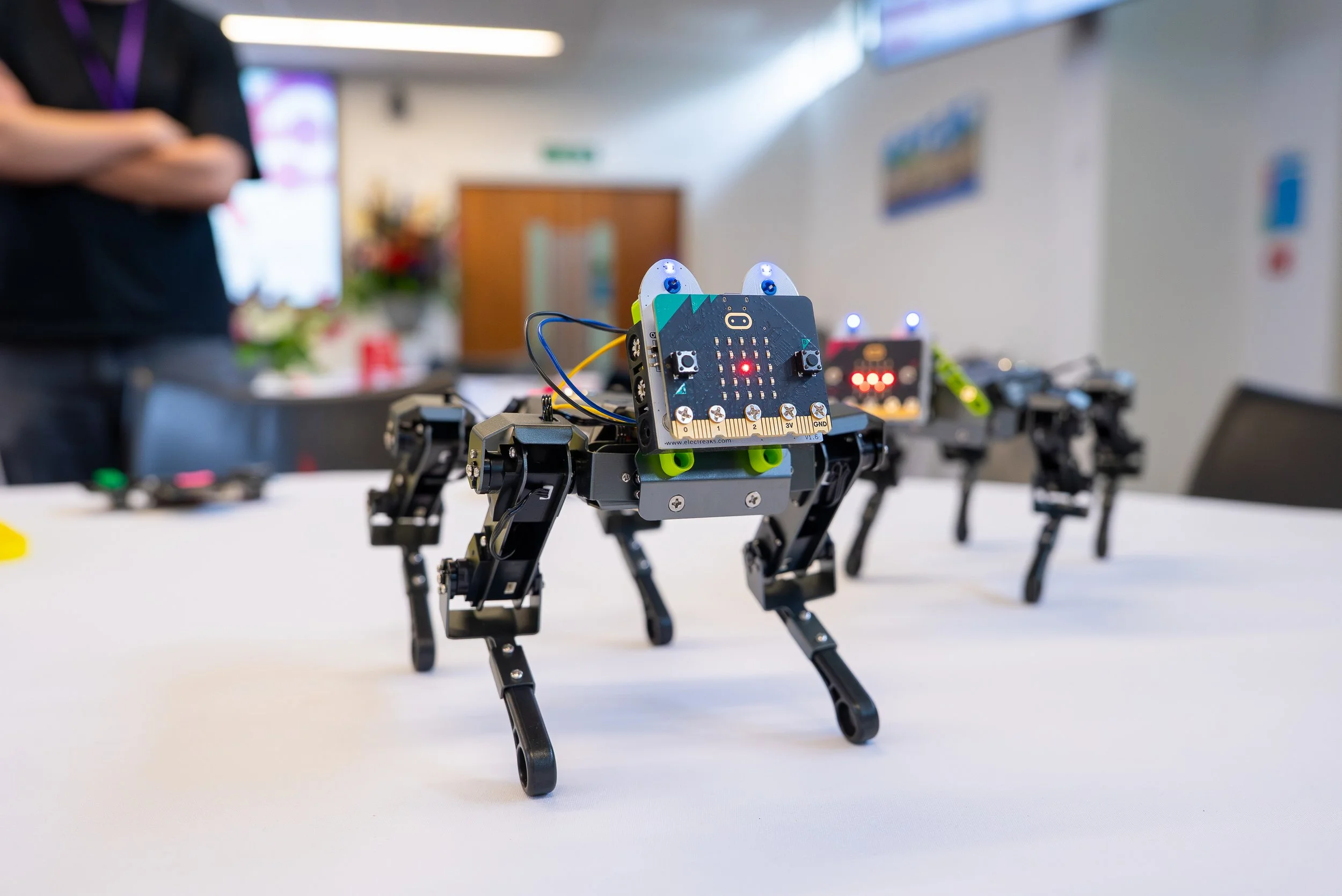 Close-up of a robot dog with a black circuit board face and glowing red and blue LEDs, standing on a white table in a room with blurred background including a woman and another robot.