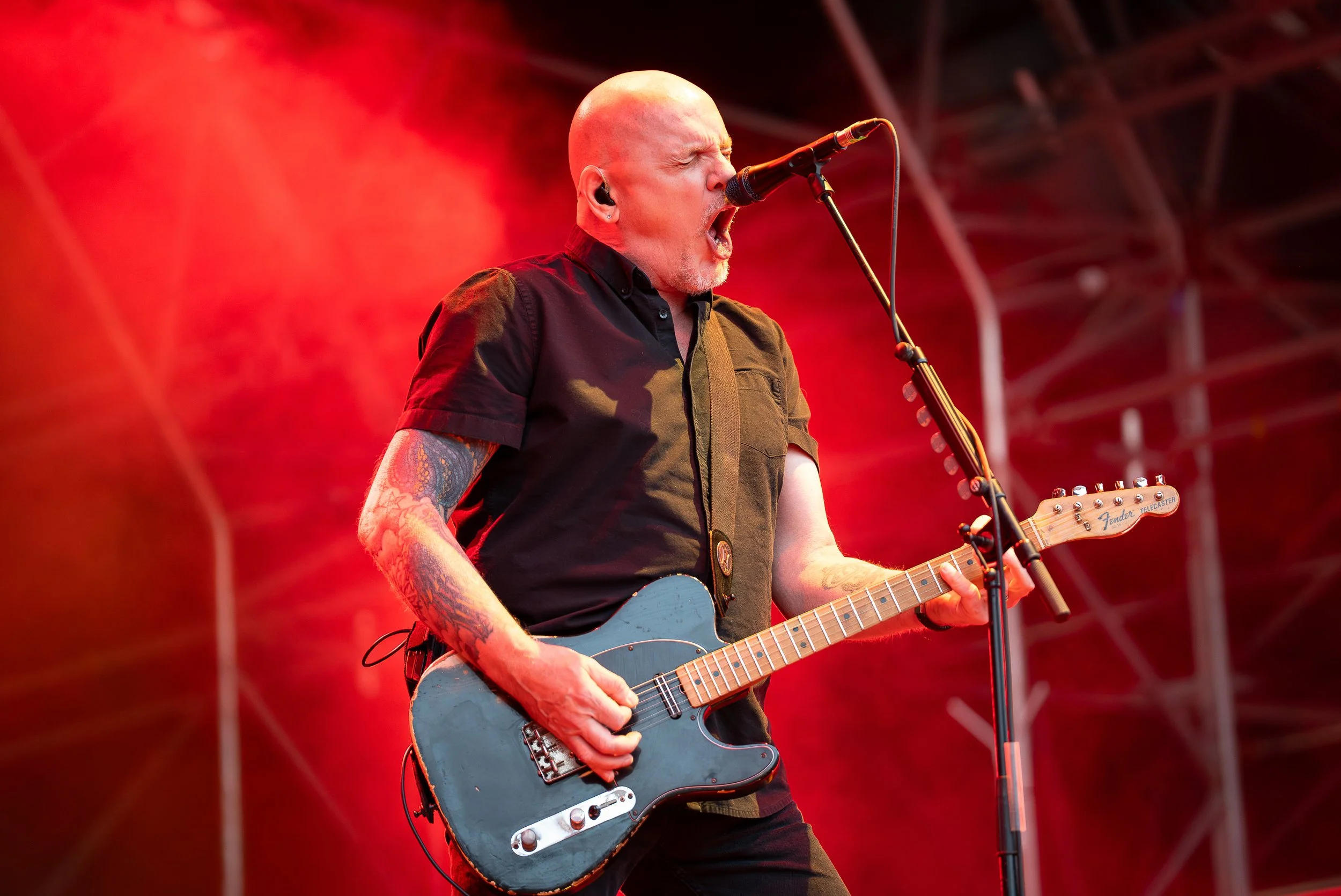 A bald man with tattoos on his arms passionately singing into a microphone while playing an electric guitar on stage, with red stage lighting in the background.