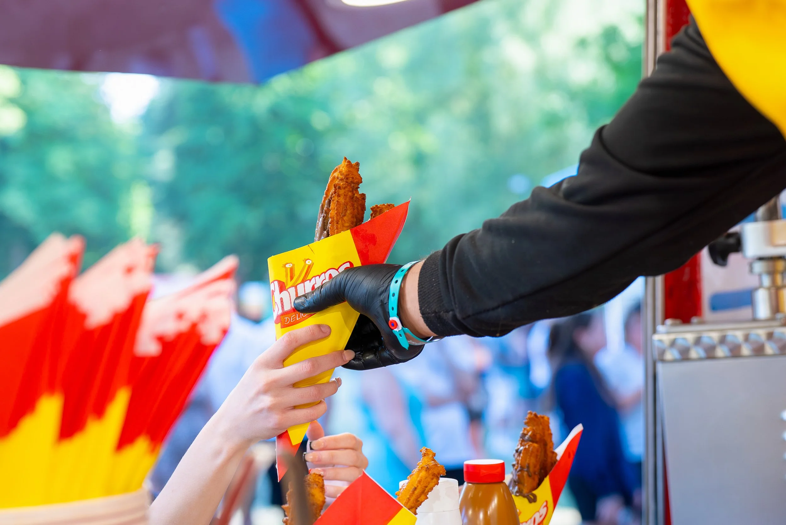 A person is handing a fried chicken tender inside a red and yellow paper cone to a child at a food stand, with bottles of dipping sauces and more chicken tenders visible on the counter.