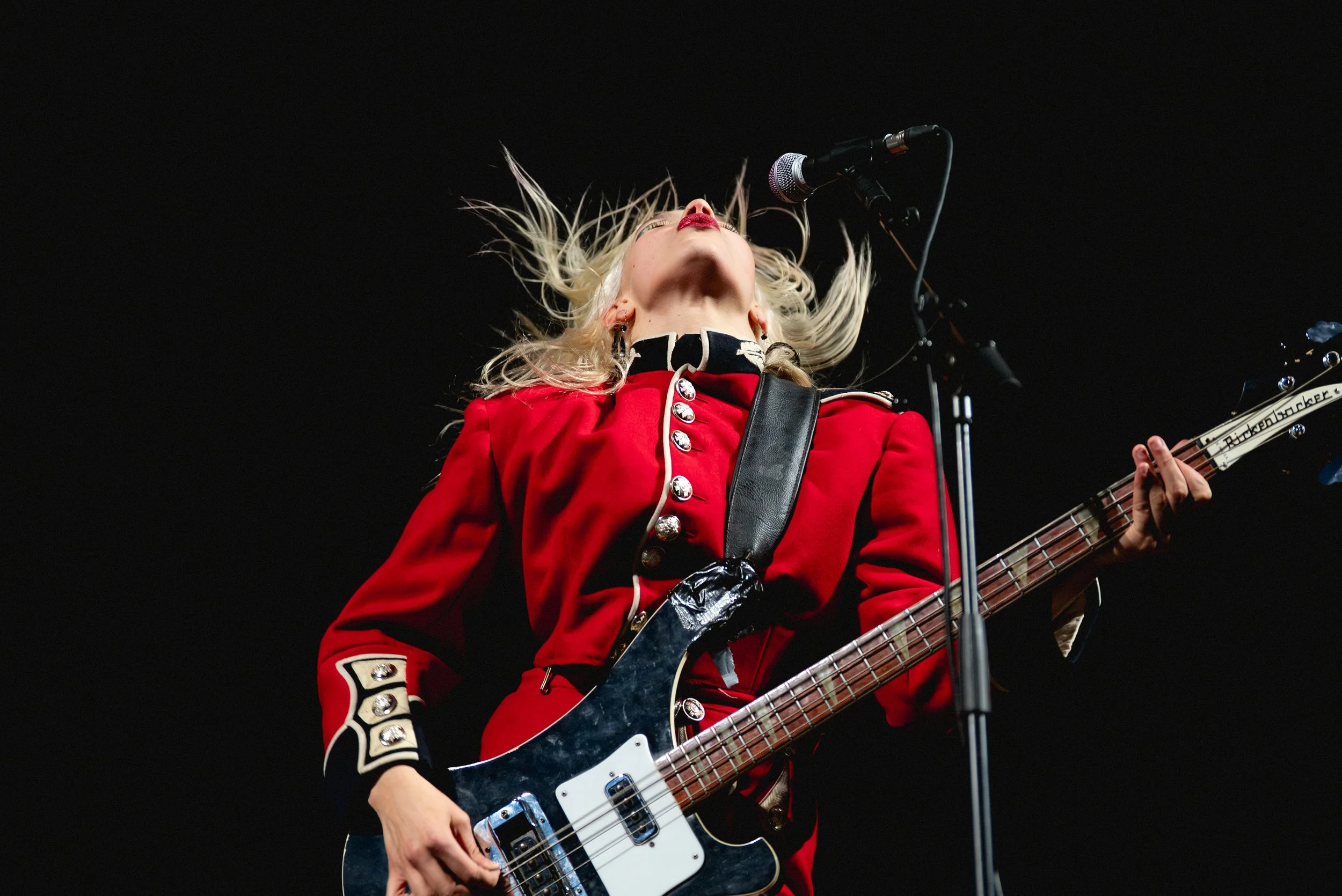 A female musician with blonde hair, wearing a red military-style jacket, playing a black and white electric bass guitar on stage. She is singing into a microphone against a black background.