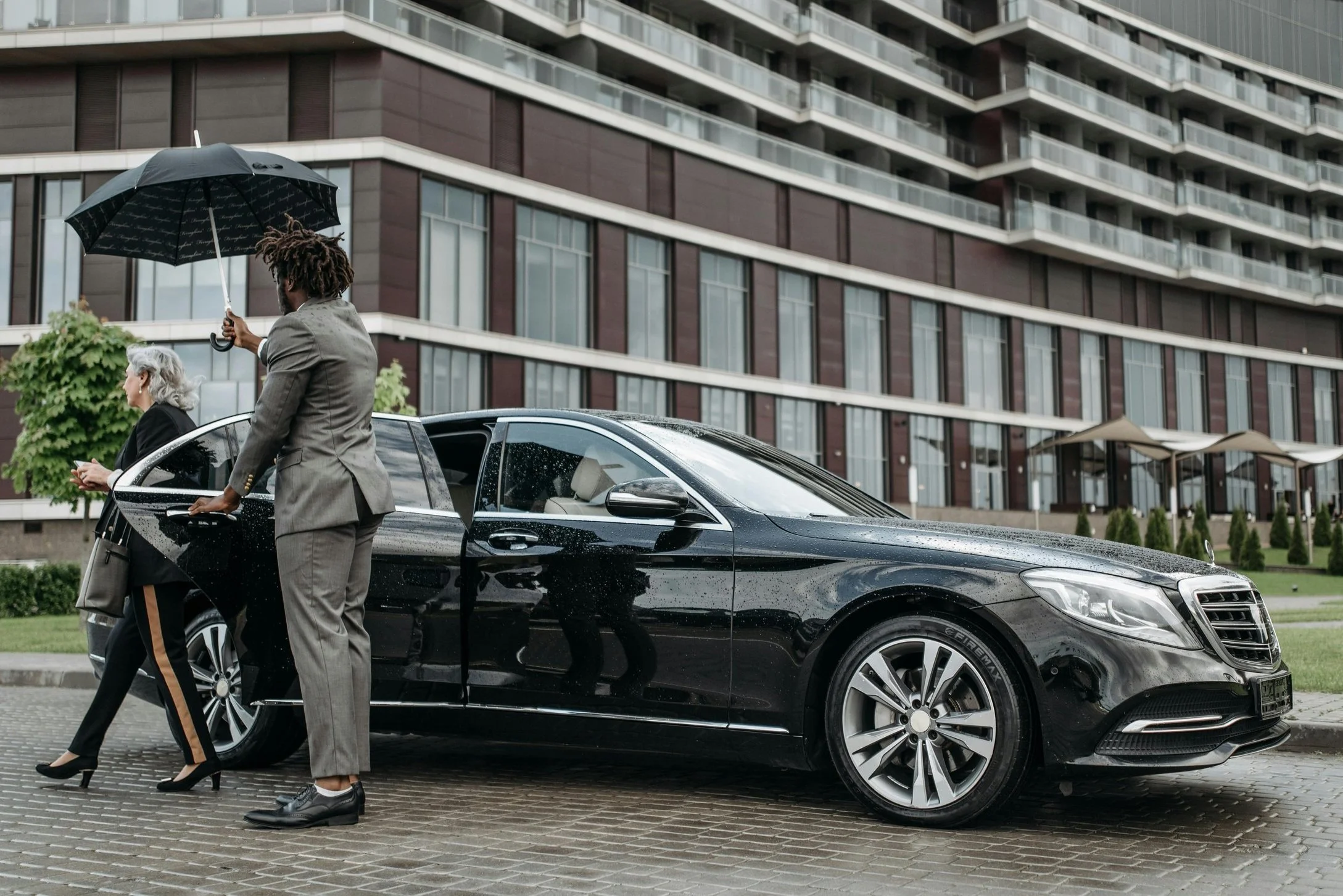Two people, one elderly woman with gray hair and a young chauffeur with dreadlocks, standing next to a black luxury car in front of a modern apartment building on a rainy day. The driver is holding an umbrella.