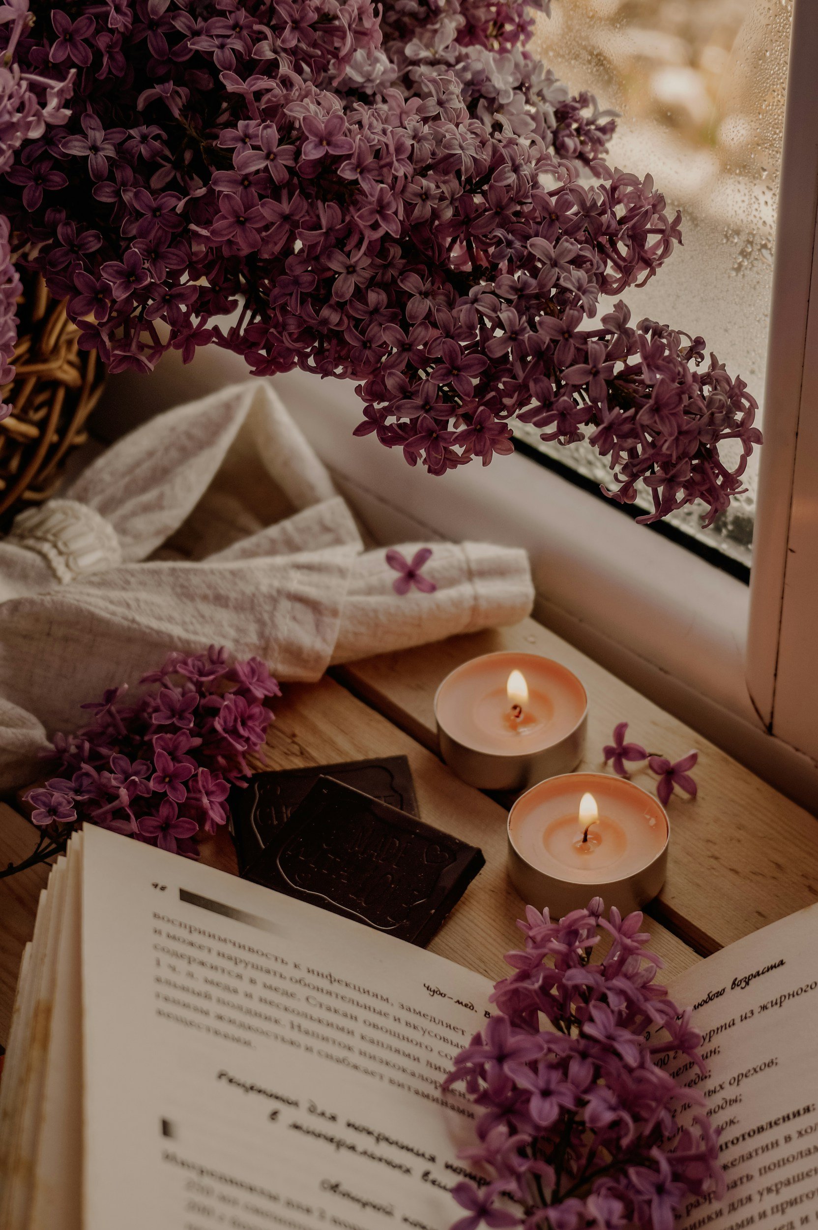 A cozy scene with purple lilac flowers, two lit candles, an open book with text in Russian, and a white cloth on a wooden surface near a window with condensation.