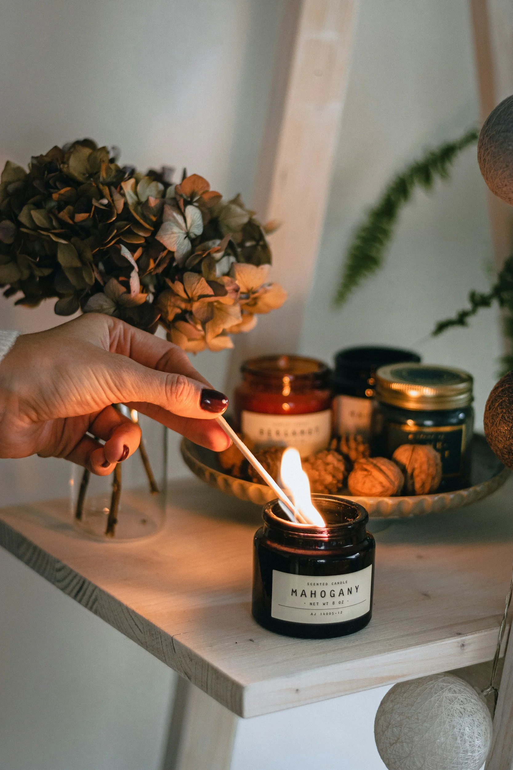 A person lights a scented candle labeled 'Mahogany' with a match on a wooden table. In the background, there are jars, a tray with walnuts, and a potted plant, with a neutral-colored wall.