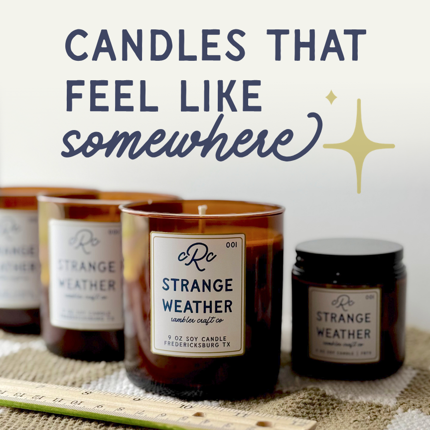 Three scented candles labeled 'Strange Weather' from Rumble Craft Co., placed on a cloth surface with a ruler in front. A sign in the background reads 'Candles that feel like somewhere' with a star graphic.