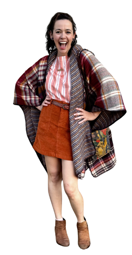 A woman with dark hair, wearing a red and white striped shirt, orange skirt, tan ankle boots, and a large plaid coat with a colorful lining, posing with a big smile and hands on hips.