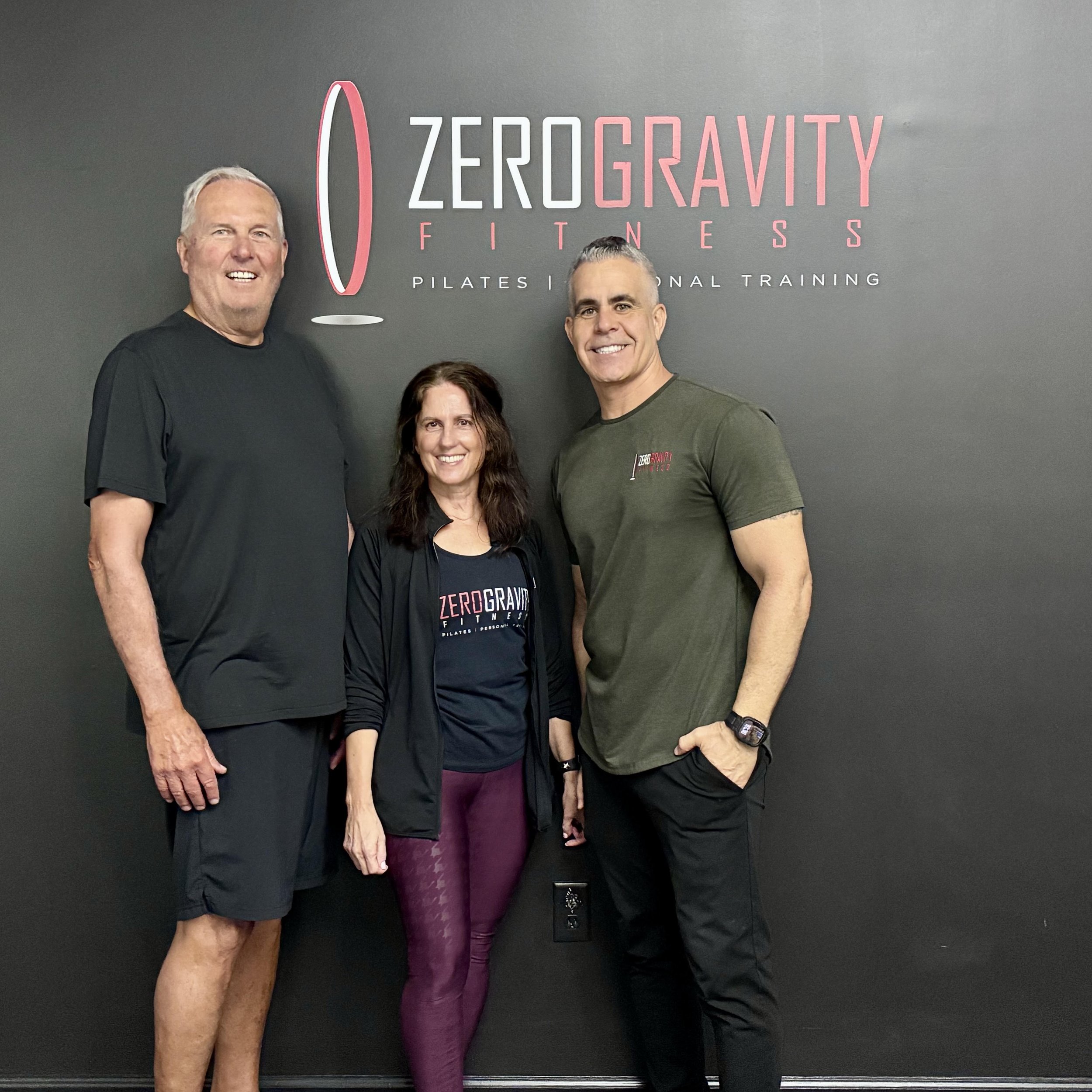 Four people standing together in front of a gray wall with 'Zero Gravity Fitness' logo, smiling.
