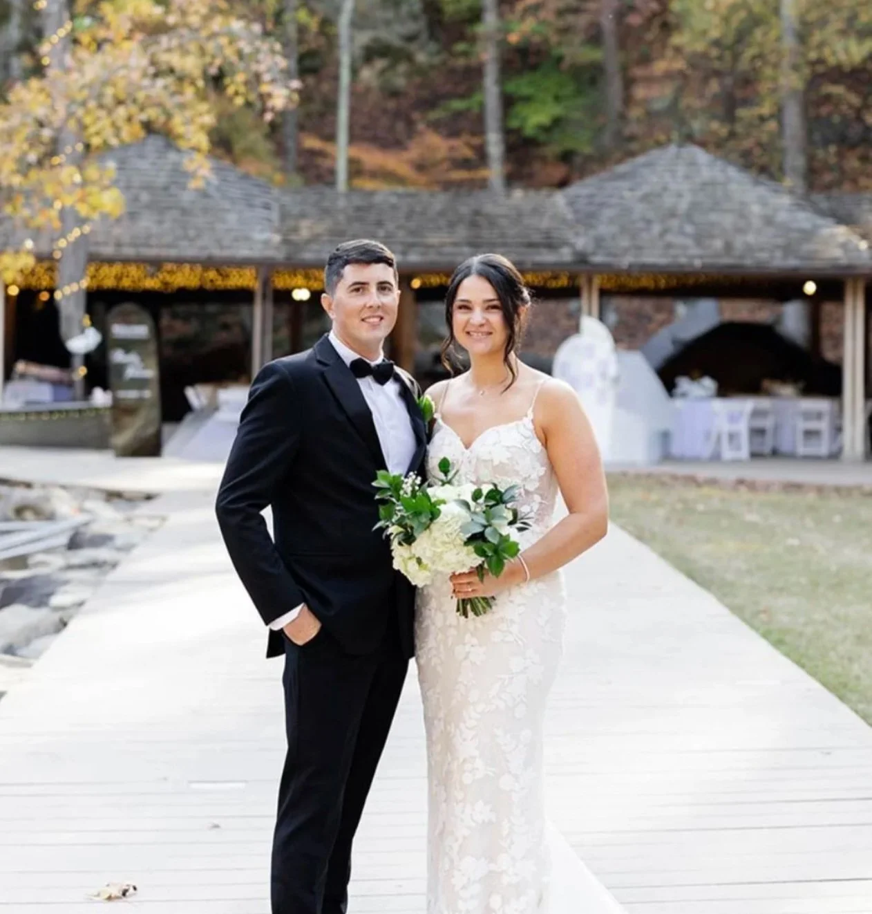 A newlywed couple at their wedding, standing close together, with the bride holding a bouquet of white and blush flowers, outdoor wedding decor with white flowers and candles in the background.