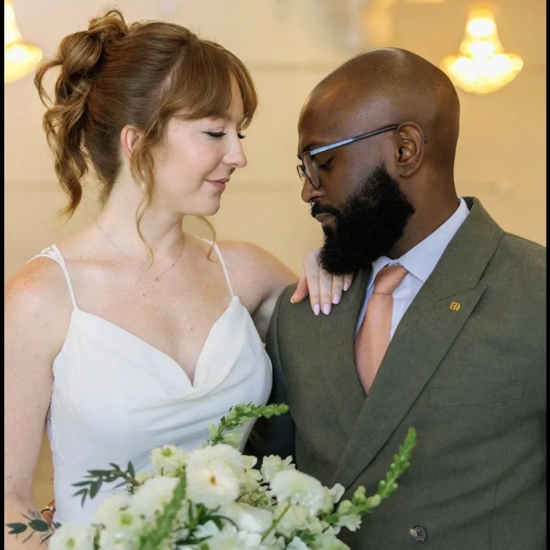A bride and groom close together in a room with wooden walls and white curtains, smiling and looking into each other's eyes.