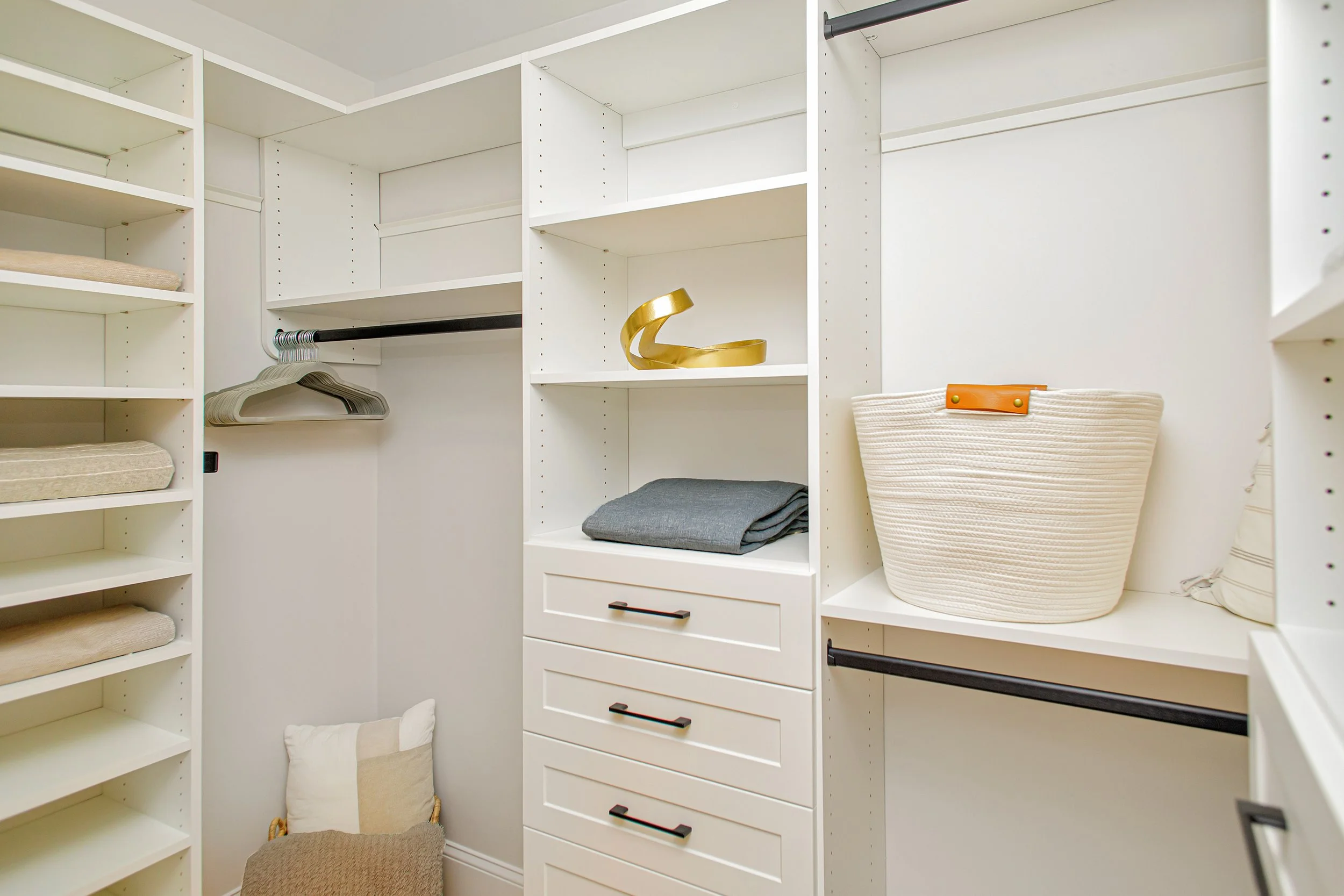 Empty white walk-in closet with shelves, drawers, and hanging rods, containing folded clothes, a gold decorative object, a large white woven basket, and a pillow and blanket on the floor.
