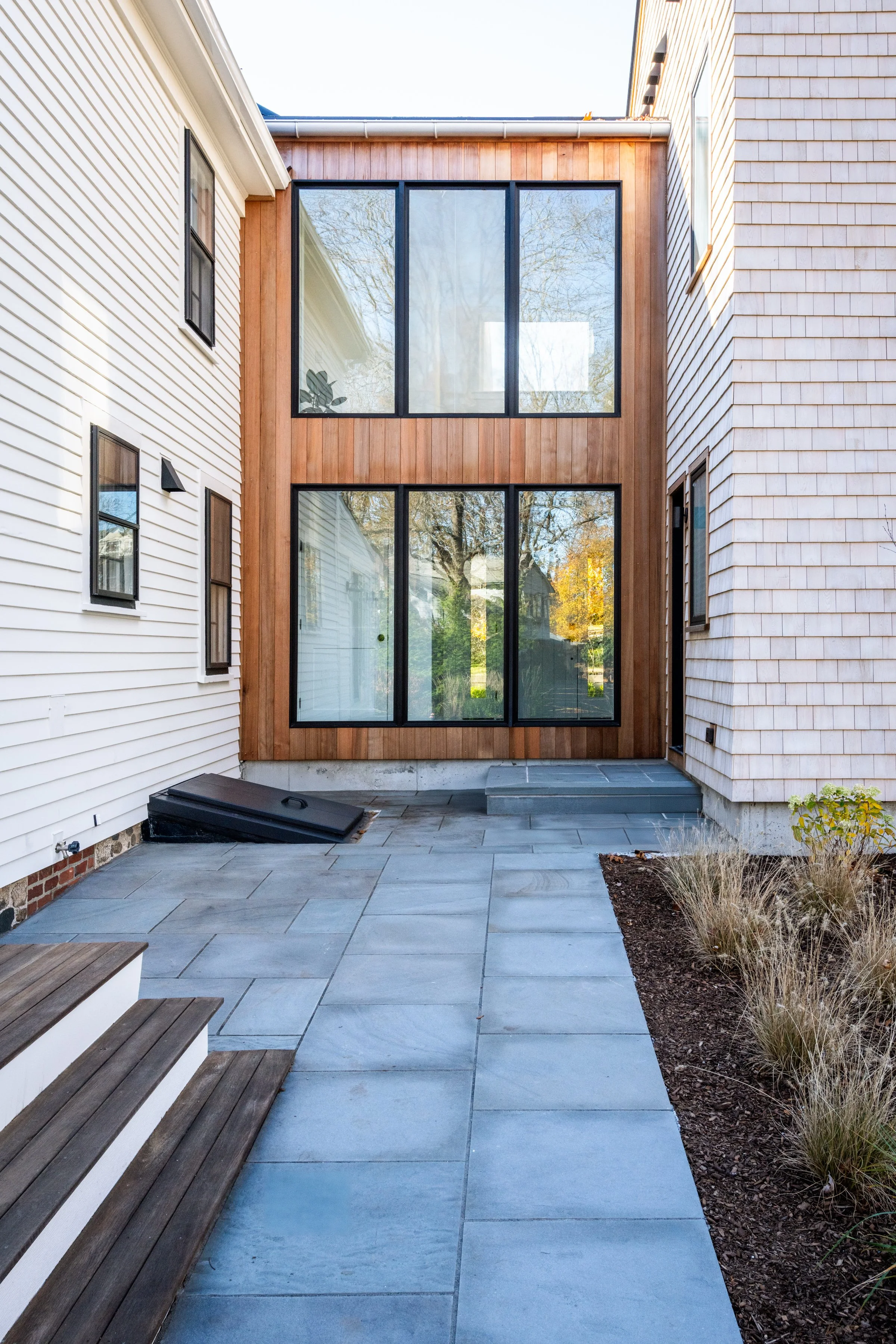 A modern residential building in Hingham, Massachusetts exterior with large glass windows, white and wood siding, three small steps leading up to a door, and a stone-paved patio with some landscaping.