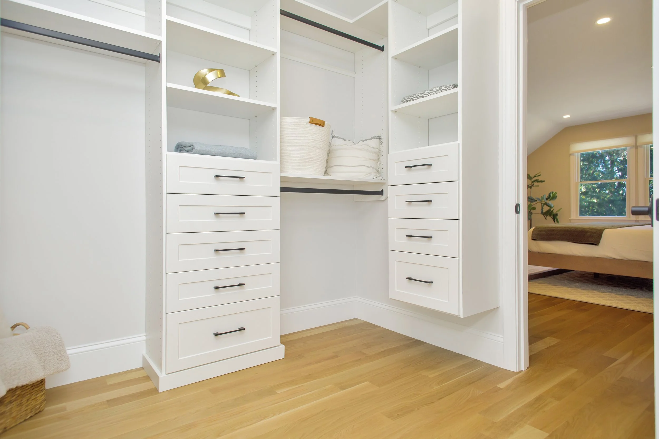 Empty walk-in closet with white shelving, drawers, and hanging rods, with a view into a bedroom with a bed and windows.