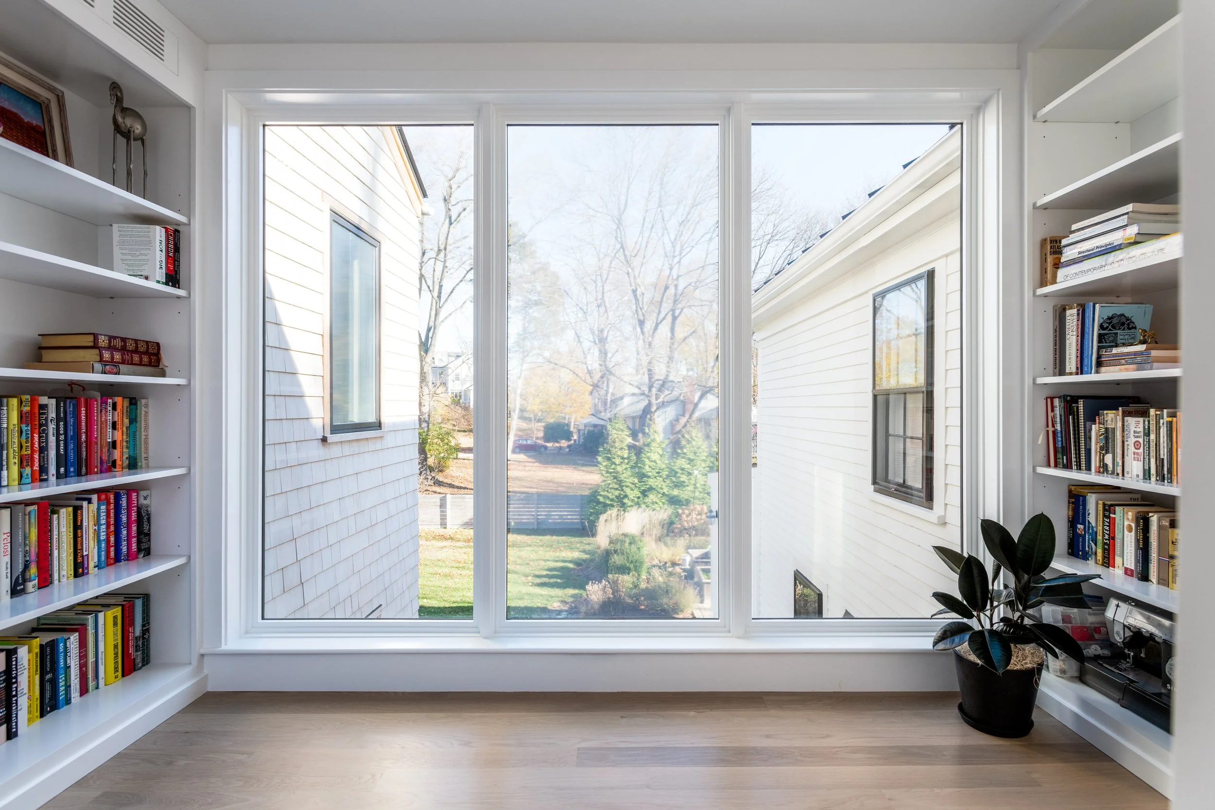 A bright room in Hingham, Massachusetts with a large window showing a view of trees and neighboring houses. The room has white bookshelves filled with books and a potted plant in the corner.