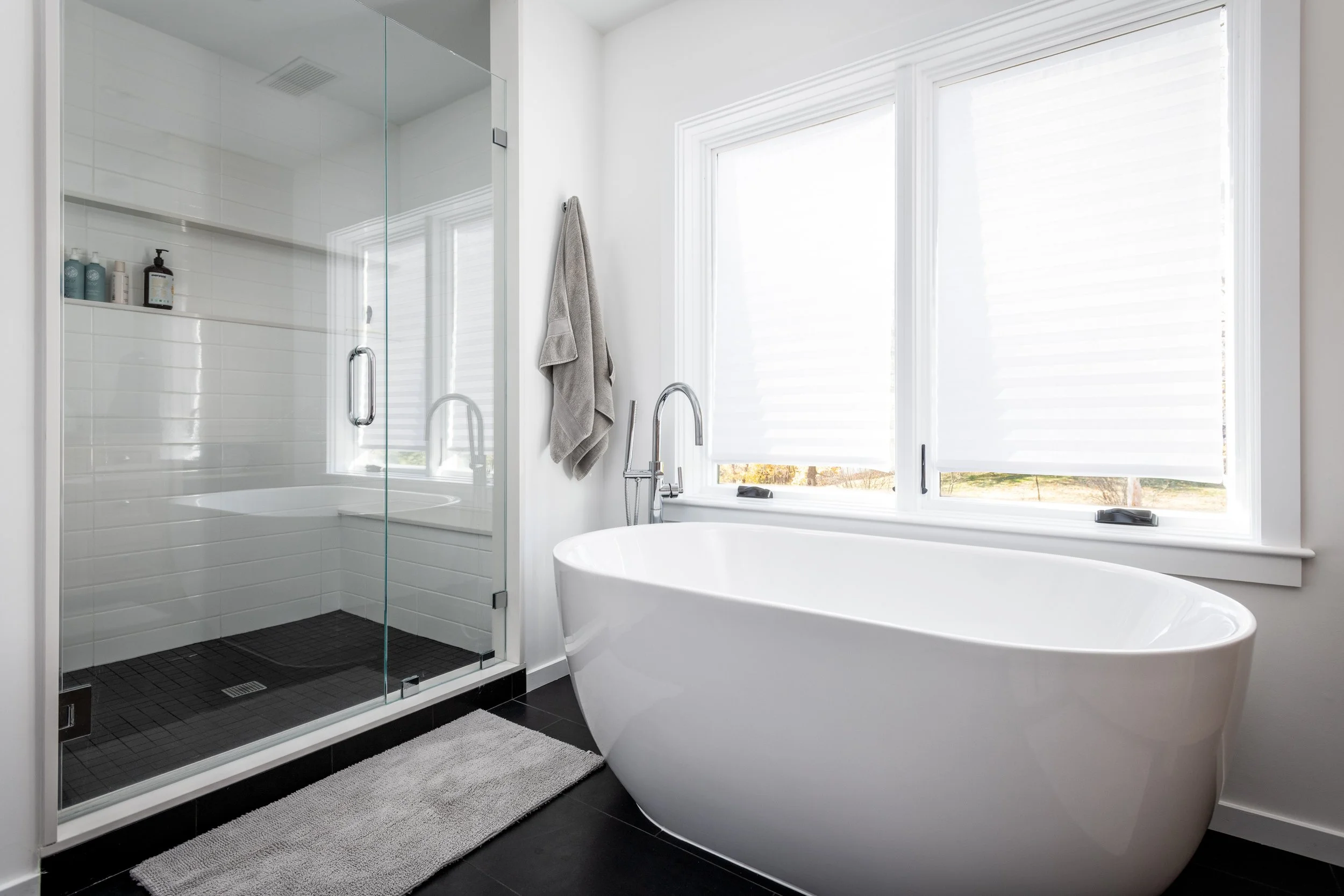 Modern bathroom in Hingham, Massachusetts with white bathtub, two large windows with blinds, gray towel hanging, shower area with glass door, black floor tiles, and a gray bath mat.