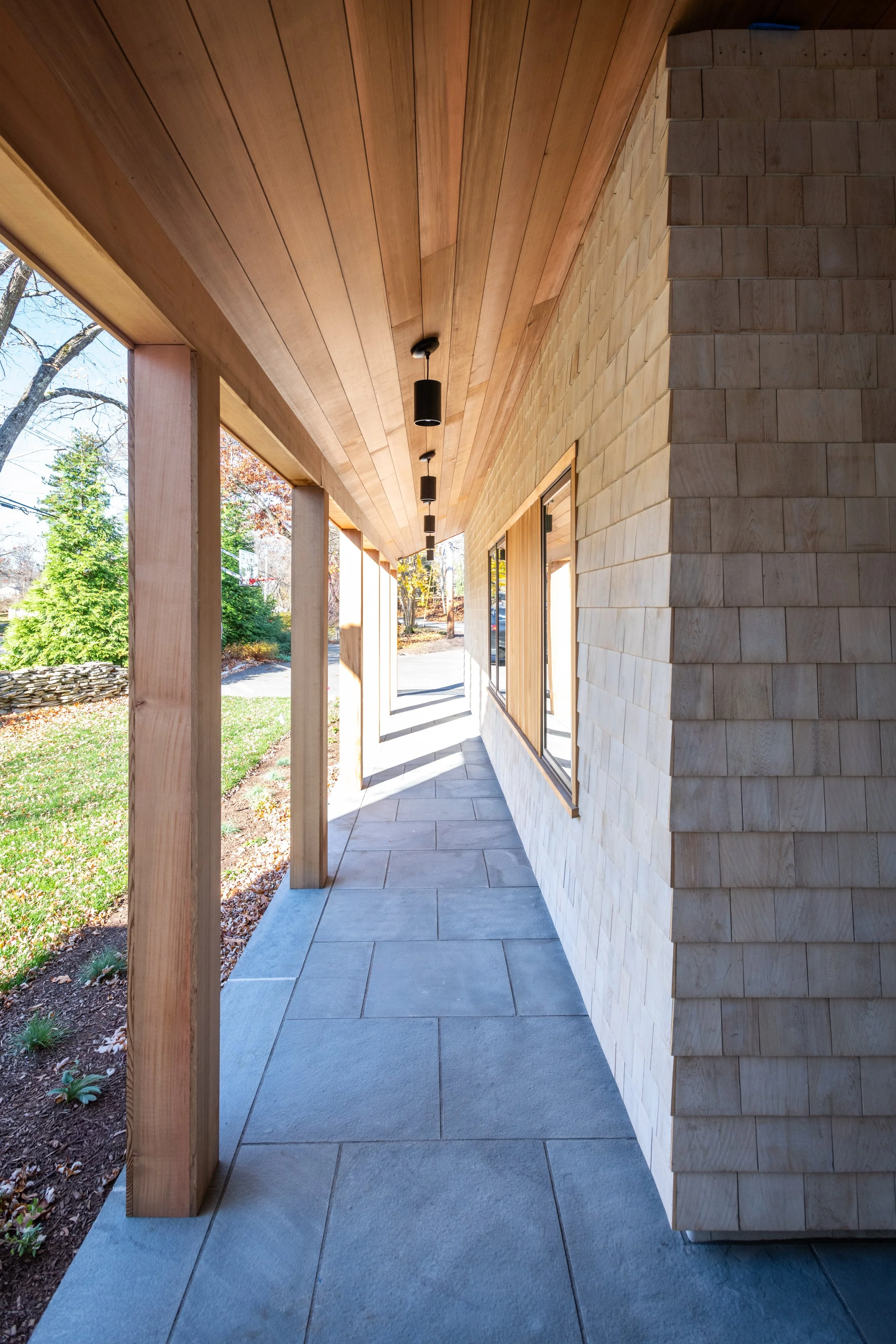 Exterior view of a modern home in Hingham, Massachusetts with a covered porch, wooden ceiling, brick siding, large windows, and pathway.
