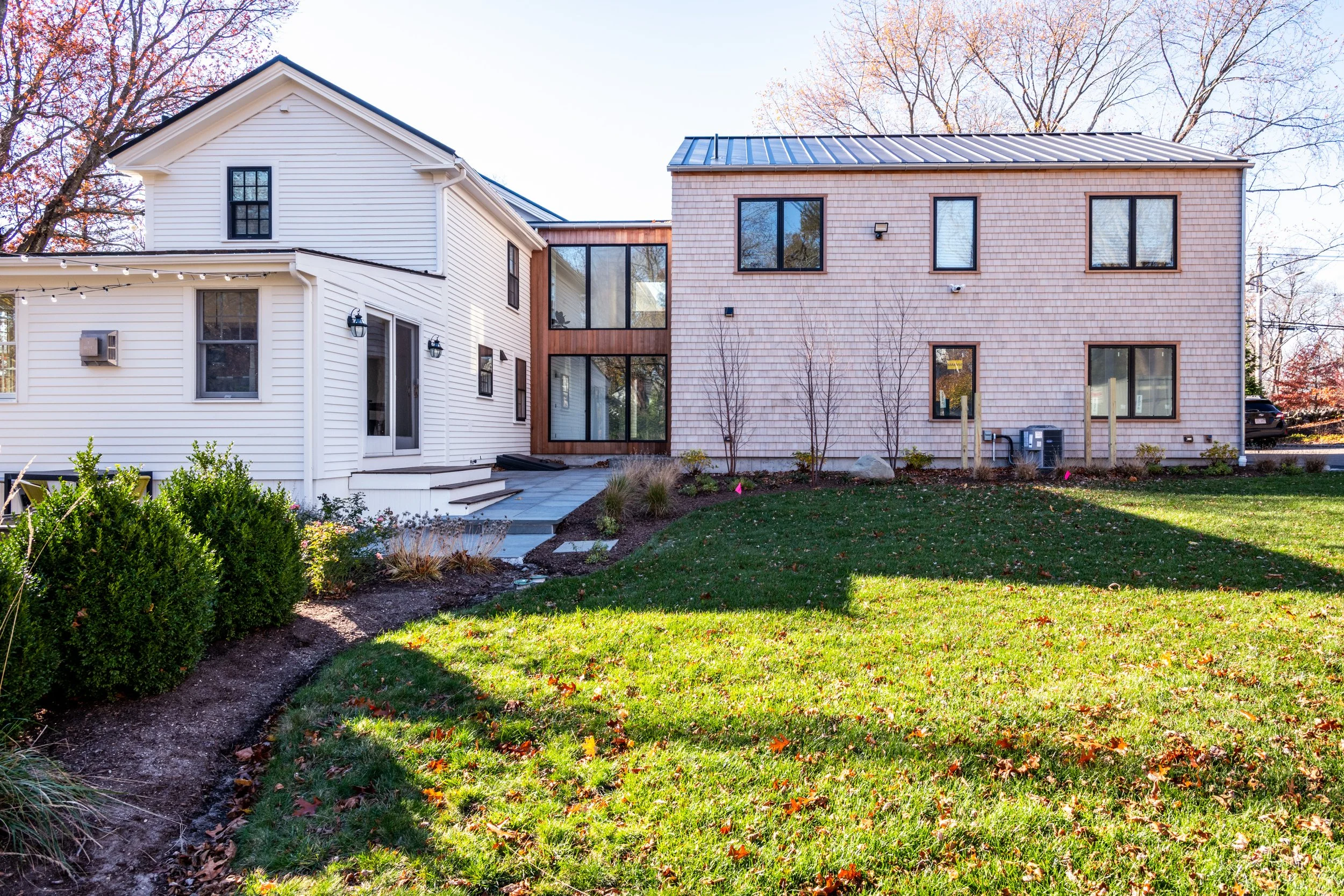 A modern home addition in Hingham, Massachusetts connected by a glass hallway, one is white with vinyl siding and the other has a tan shingle exterior, surrounded by a grassy lawn with small trees and plants.