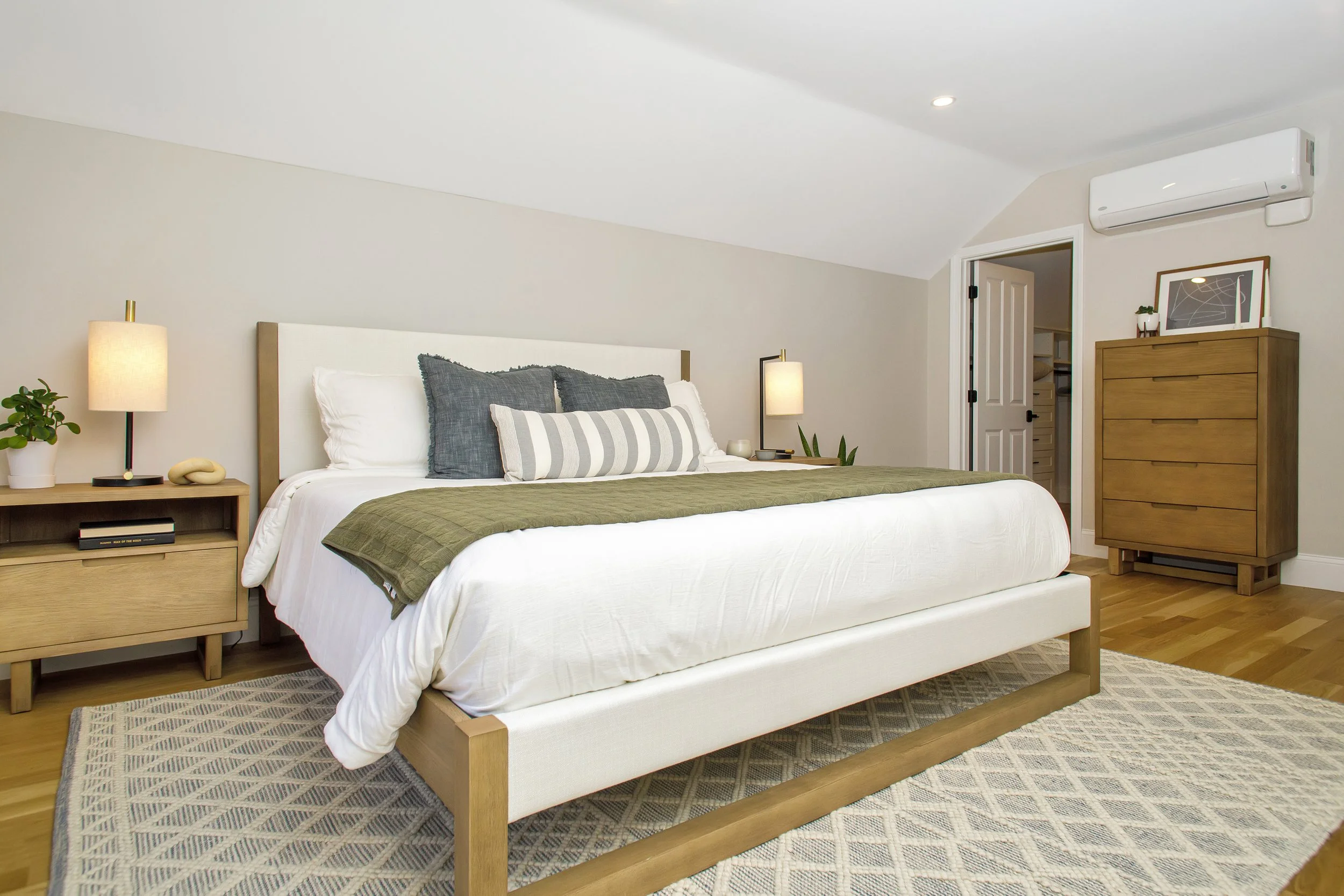A modern bedroom with a large bed, nightstands, lamps, dresser, and air conditioning unit.