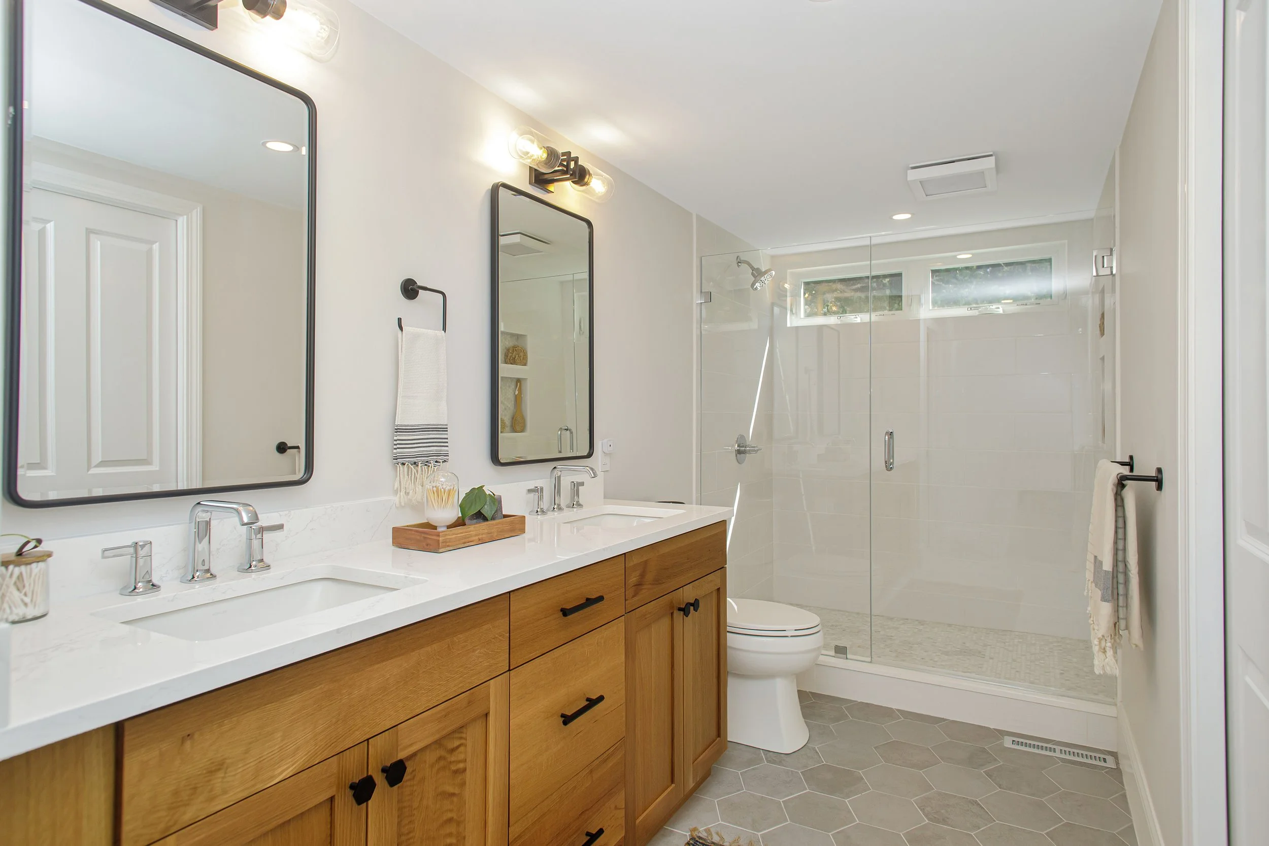 A modern bathroom featuring a double vanity with a white countertop, wooden cabinetry, and black hardware, two mirrors above the sinks, wall-mounted lighting, a toilet, and a glass-enclosed shower with a small window.