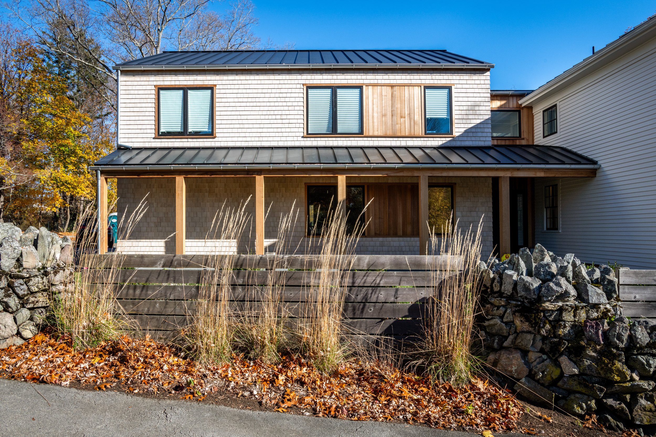 Newly constructed two-story house with a front porch, black metal roof, and mixed siding of shingles and wood paneling. The house has multiple windows with blinds, and there are trees with colorful fall leaves in the background. A stone and wooden retaining wall with grasses and fallen leaves in front.