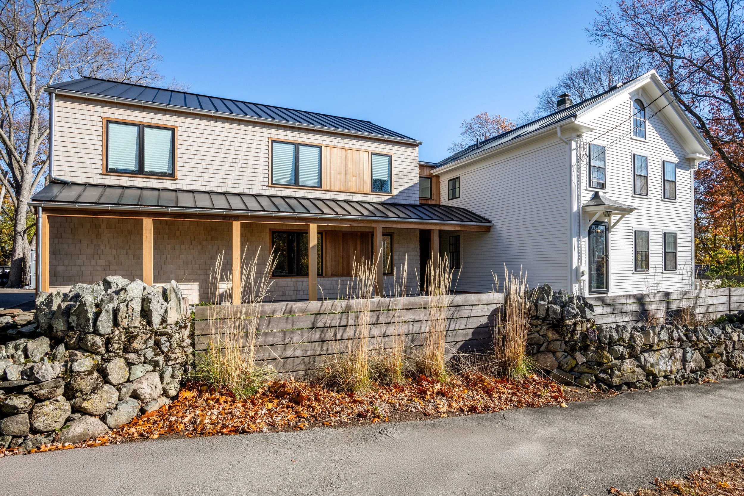 A modern house in Hingham, Massachusetts with a combination of white siding and wooden shingles, metal roof, and a small porch area, set against a backdrop of autumn trees and blue sky.