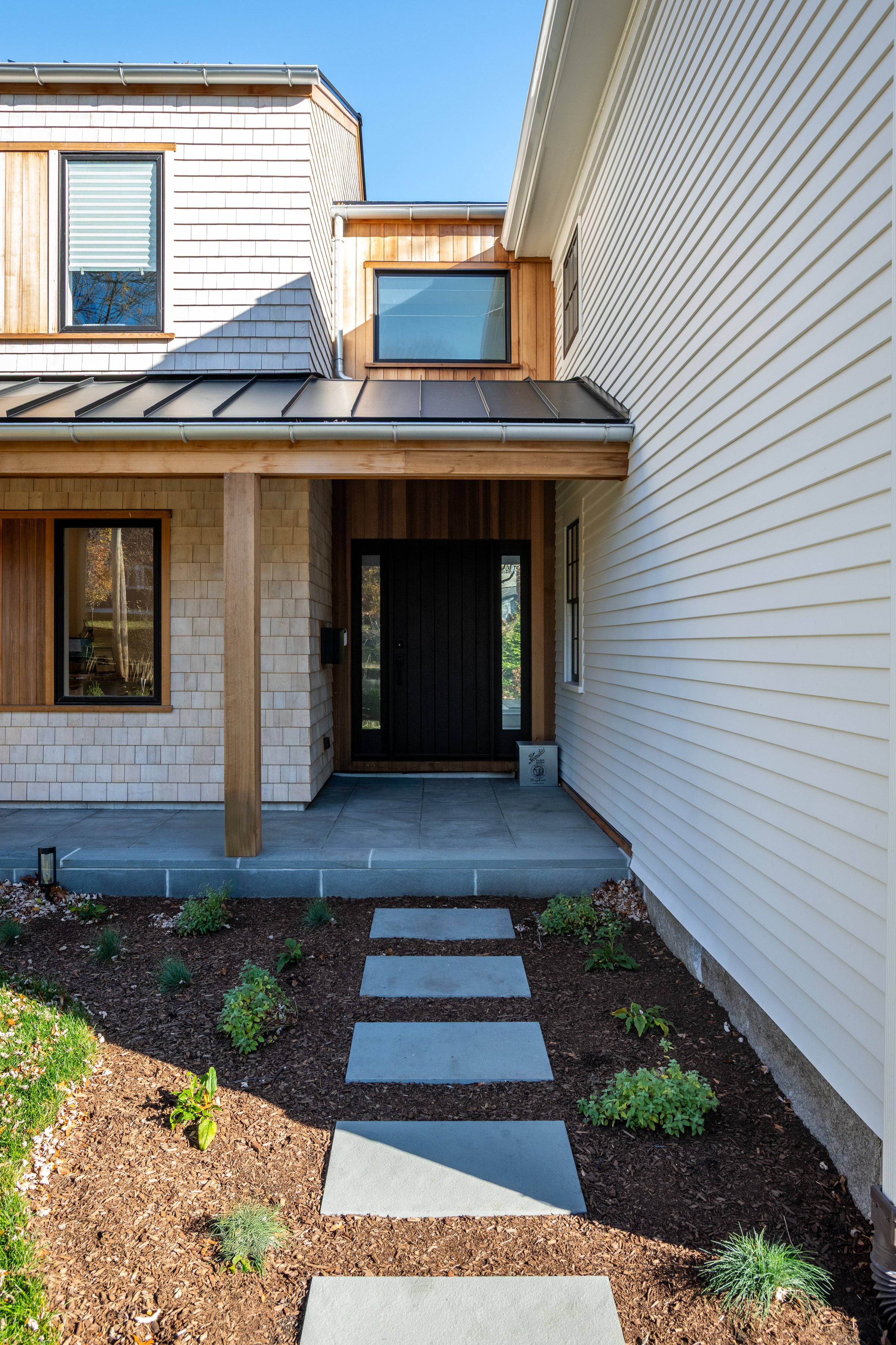 Front entrance of a modern house in Hingham, Massachusetts with a black door, stone and wood siding, and a small garden with stepping stones leading to the door.