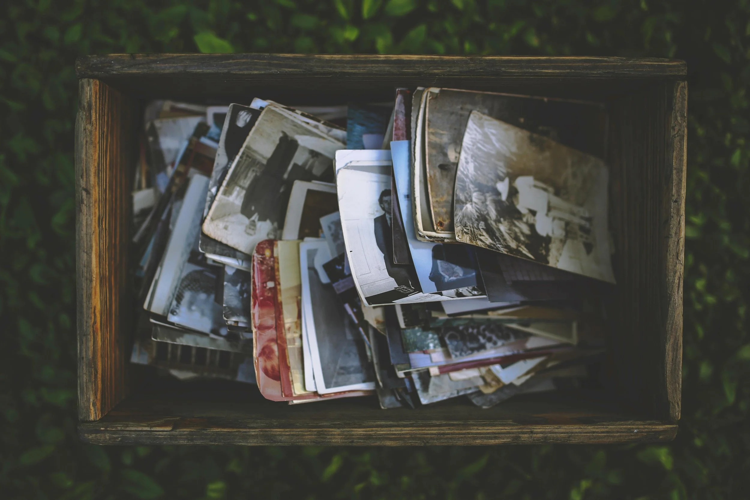 A wooden box filled with old, torn black-and-white and color photographs stacked haphazardly.