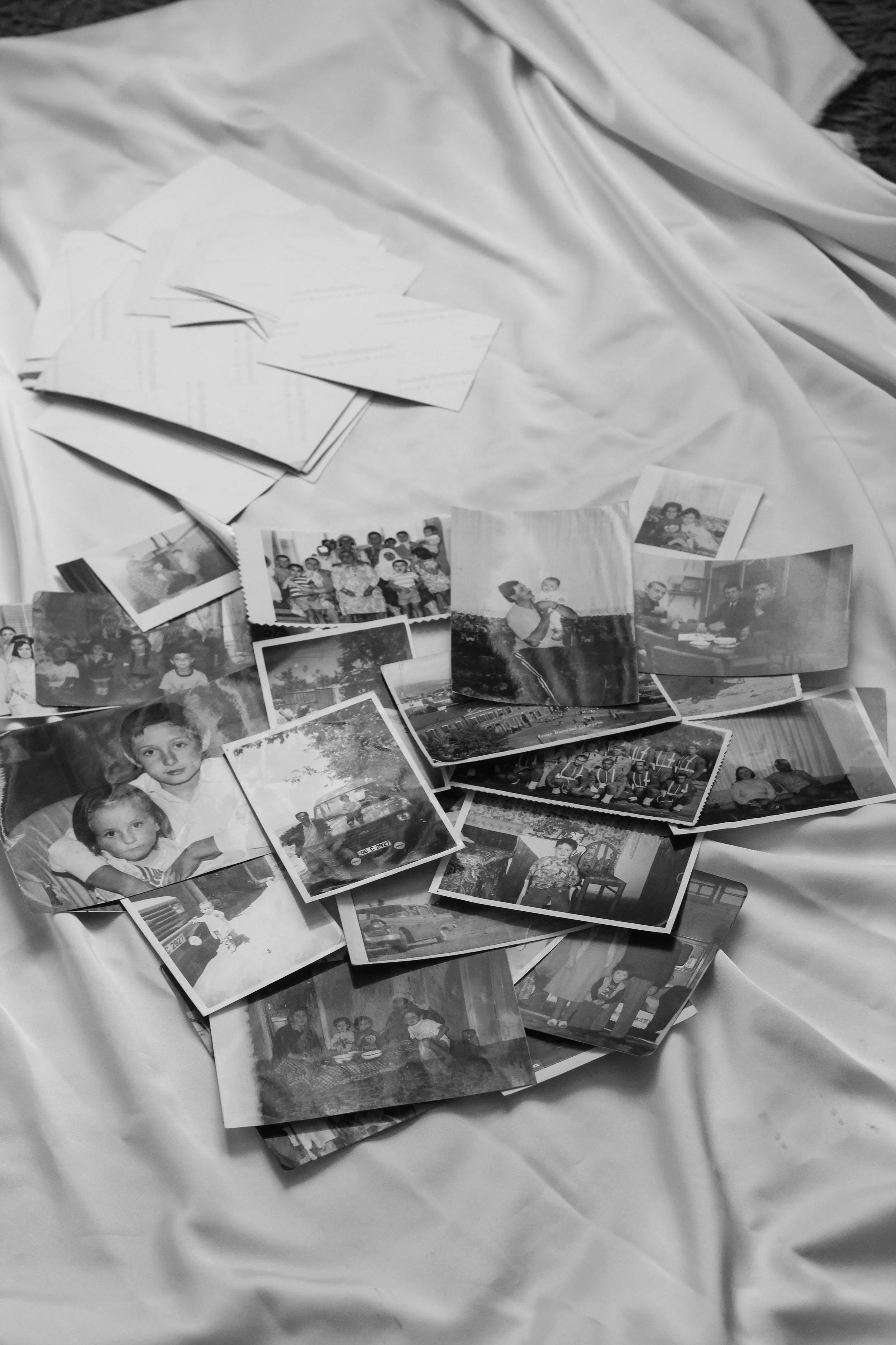 Black and white photo of scattered old photographs and envelopes on a silky cloth surface, depicting people, cars, and various scenes.